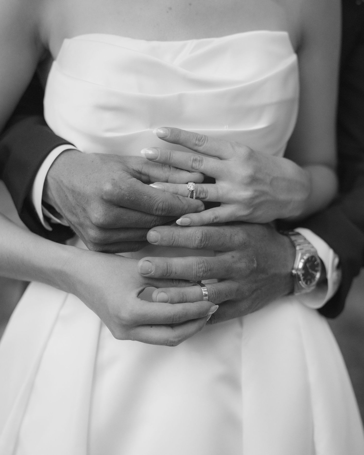 Black and white photo of newlyweds hands with wedding rings, bride in strapless gown, groom in suit with watch.