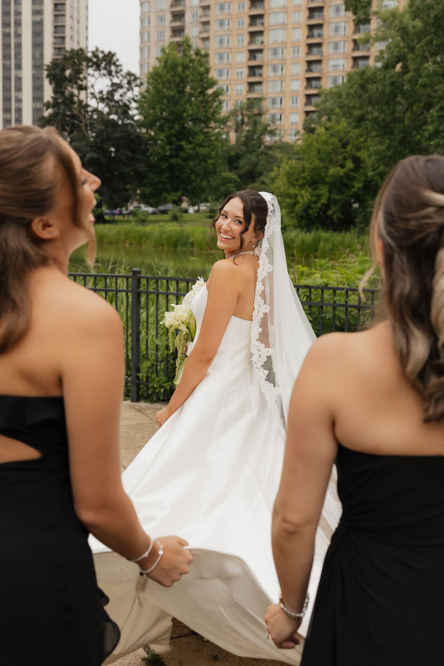 Smiling bride in white gown and veil holds bouquet while bridesmaids in black dresses carry her train outdoors.