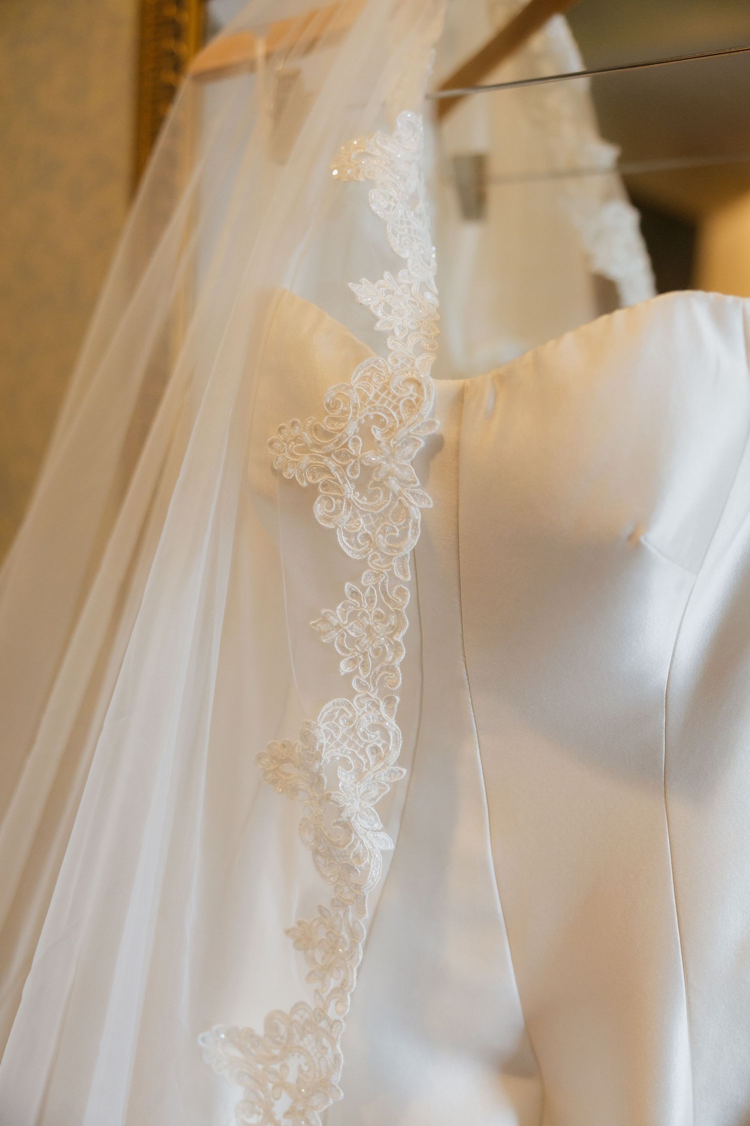 Elegant white wedding dress with lace-trimmed veil hanging on a rack, showcasing intricate floral lace detailing.