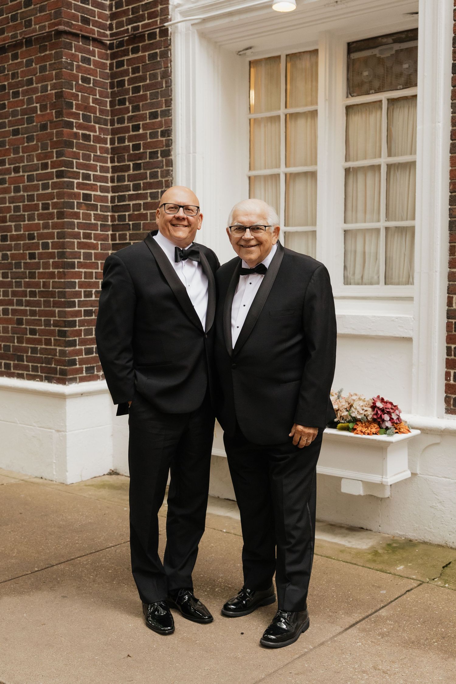Two men in black tuxedos with bow ties smiling together outside a brick building.