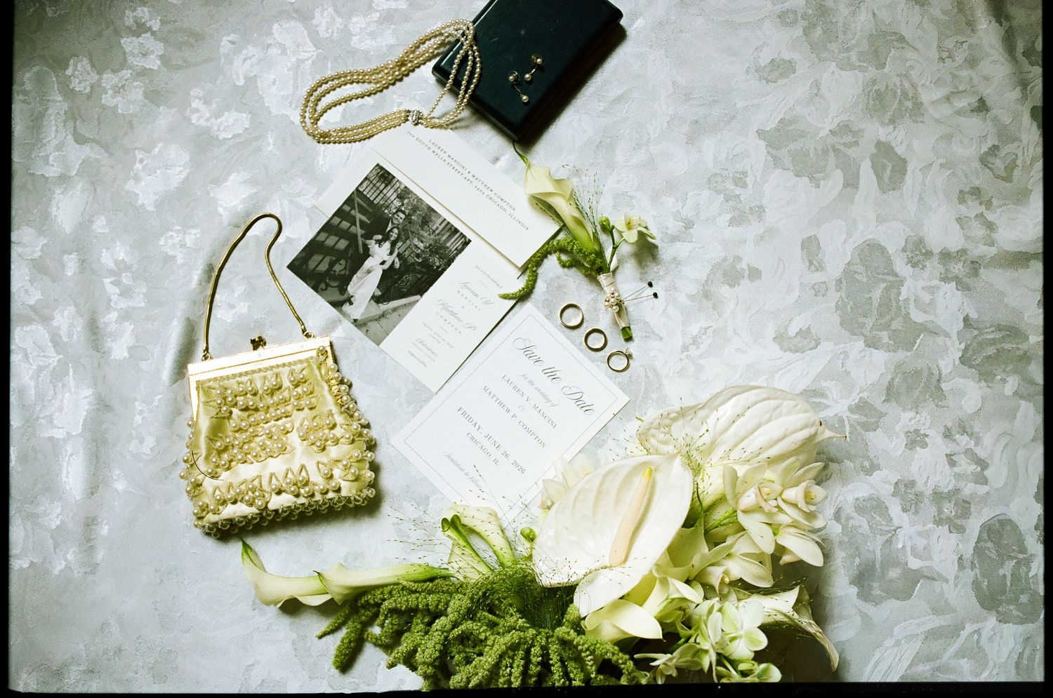 Wedding flat lay with pearl clutch, bouquet, rings, program, and black photo on floral background.