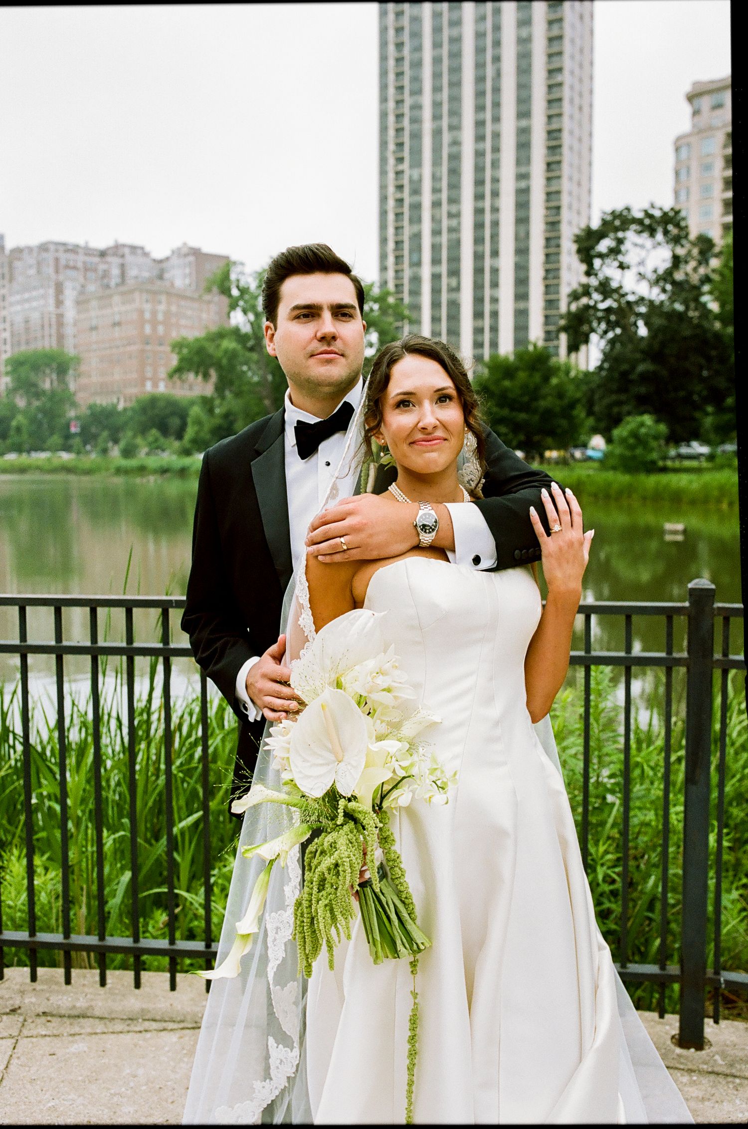 Bride and groom posing by a lake in a park, she holds white calla lilies, city skyline in background.