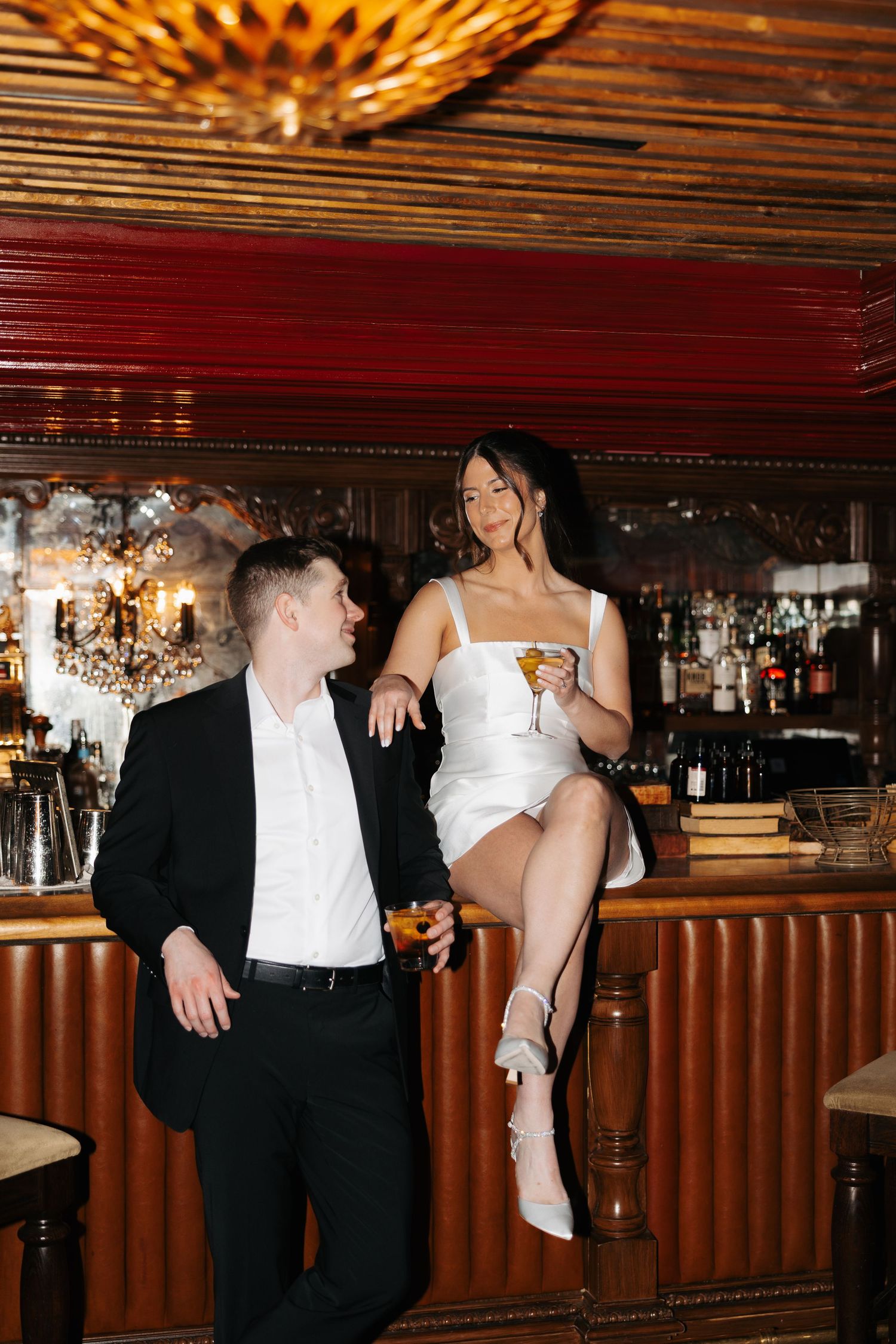 A couple in formal attire shares an intimate moment at a dimly lit bar with wood paneling and warm lighting.