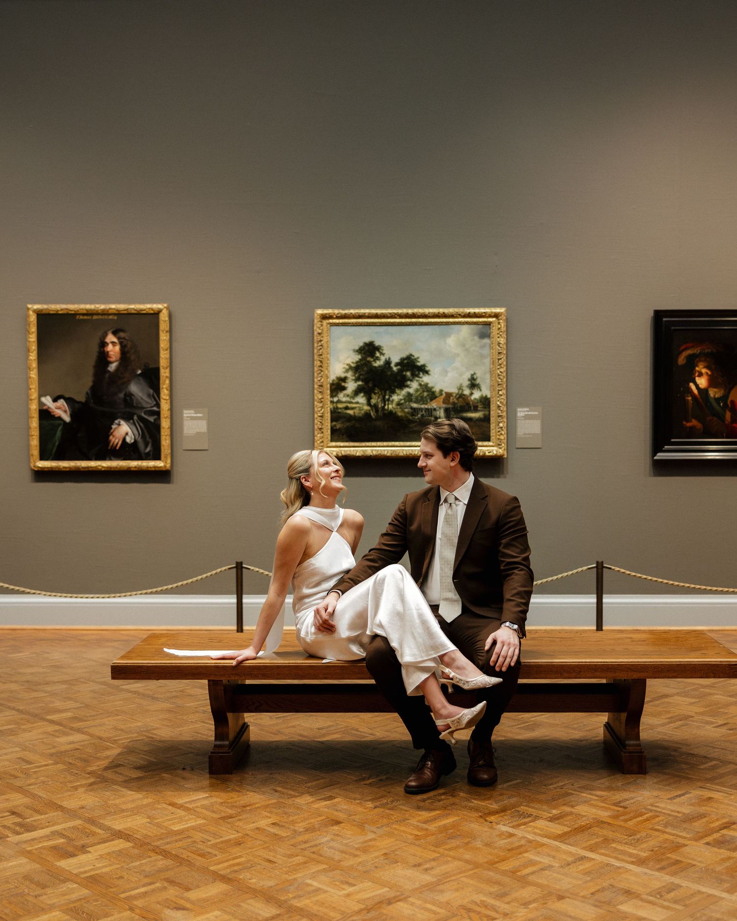 Couple in wedding attire sitting on a museum bench surrounded by classic oil paintings in an art gallery.