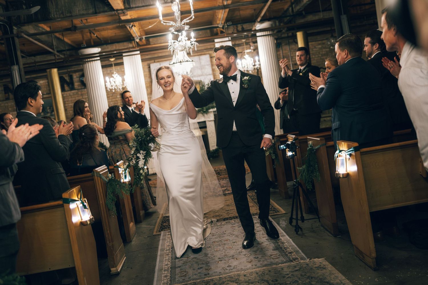 Wedding guests dance and celebrate on a dimly lit wooden dance floor during an evening reception celebration.