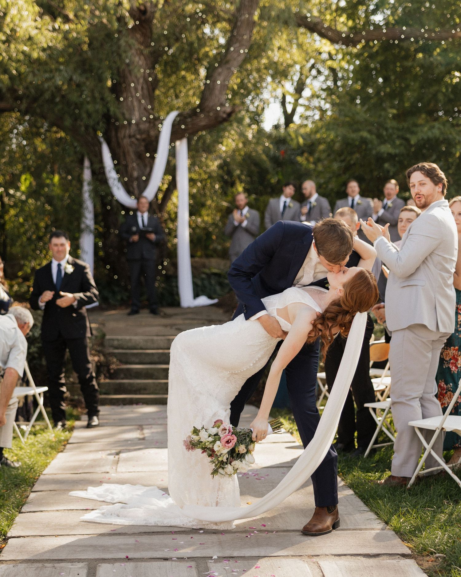 A romantic dip kiss between newlyweds at an outdoor garden wedding ceremony with guests looking on in the background.