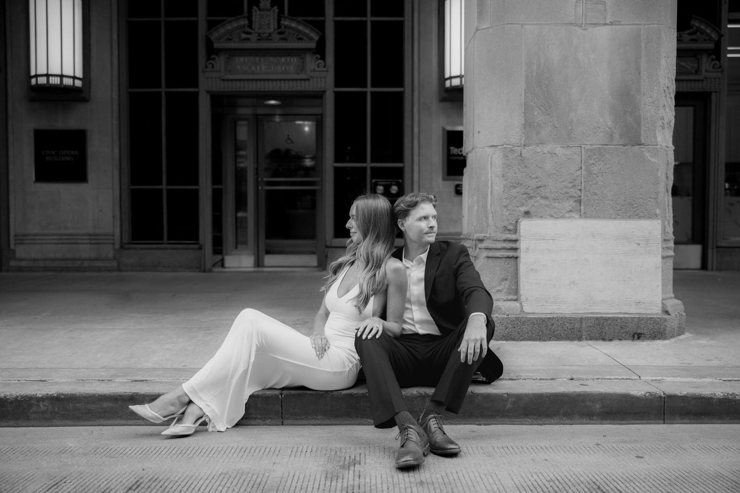 Black and white photo of a couple sitting on urban steps, woman in white pantsuit, man in dark blazer.