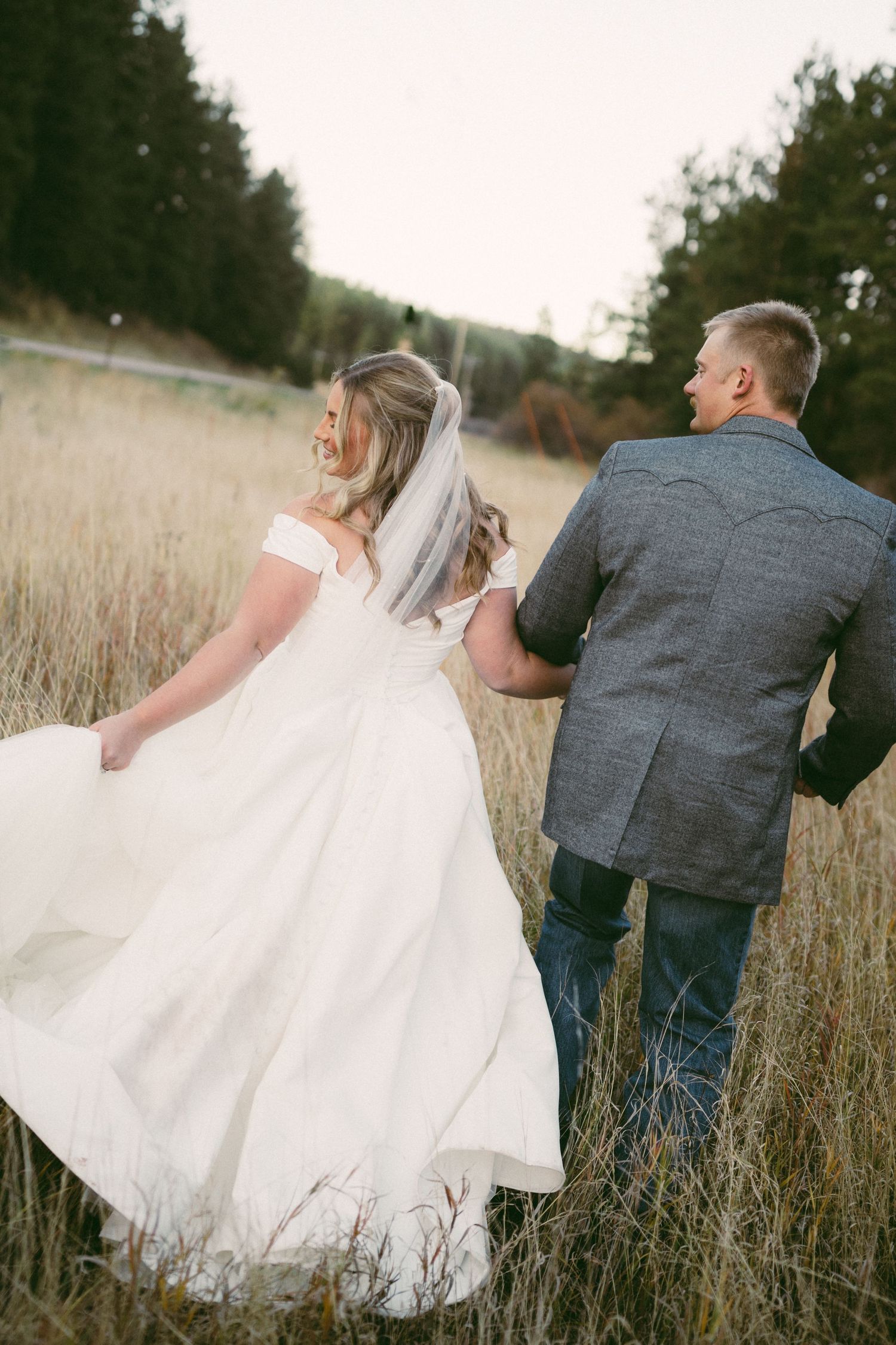 Bride in white gown and veil walks hand-in-hand with groom in gray suit through golden field.
