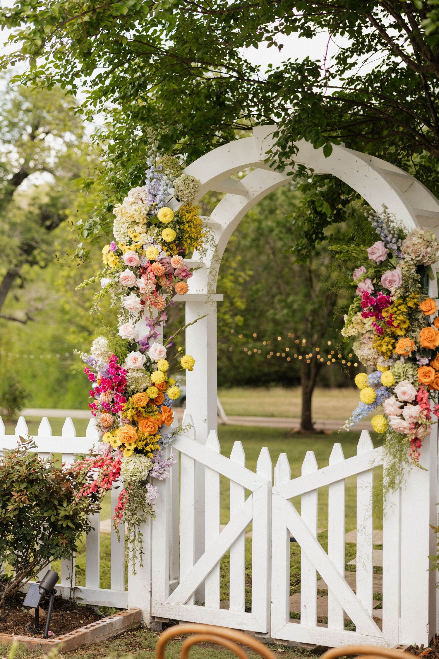 White garden arch gate adorned with colorful spring flowers including pink, yellow, and orange blooms in an outdoor setting.