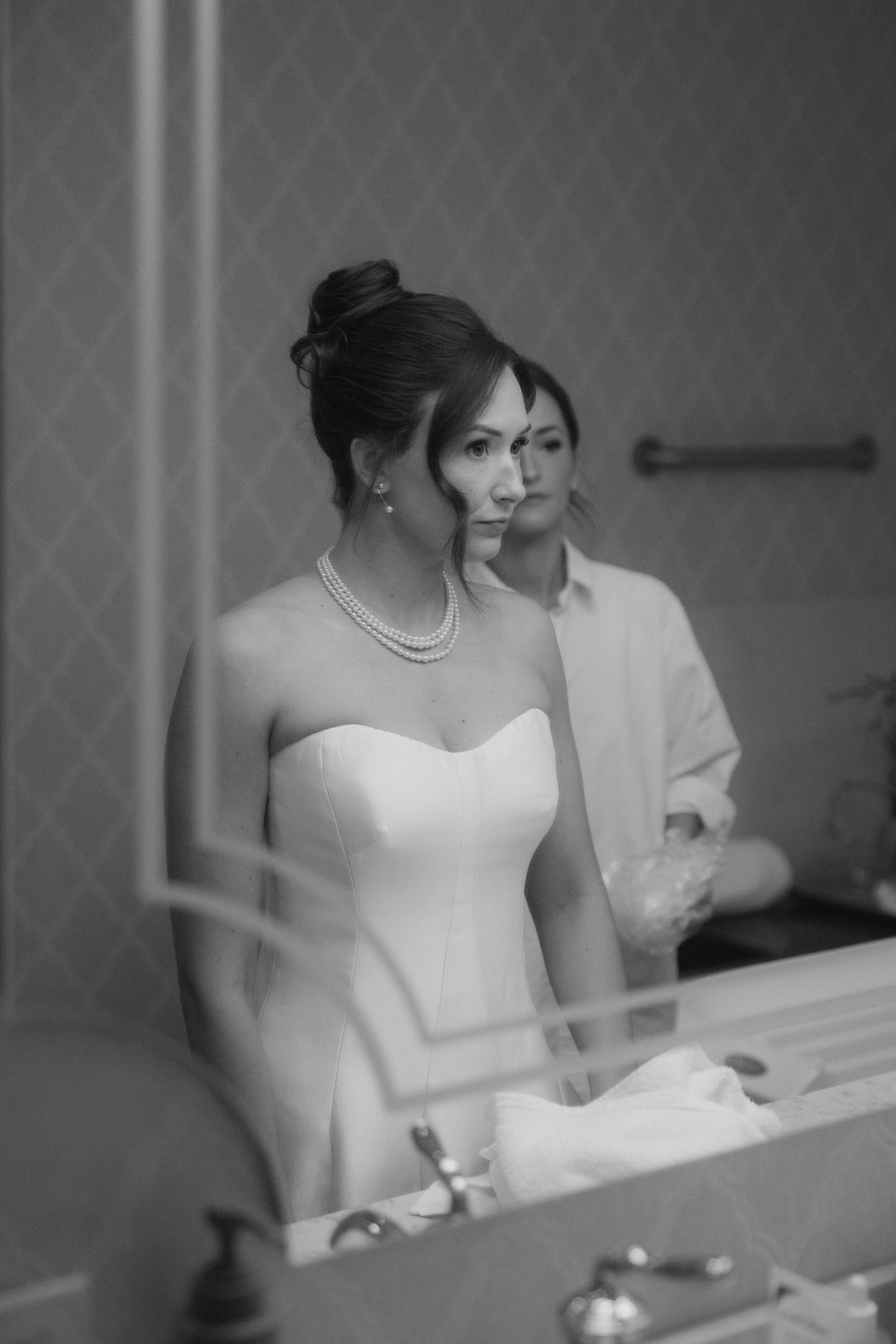Black and white photo of a bride in a strapless gown and pearl necklace reflected in a mirror as a woman stands behind her.