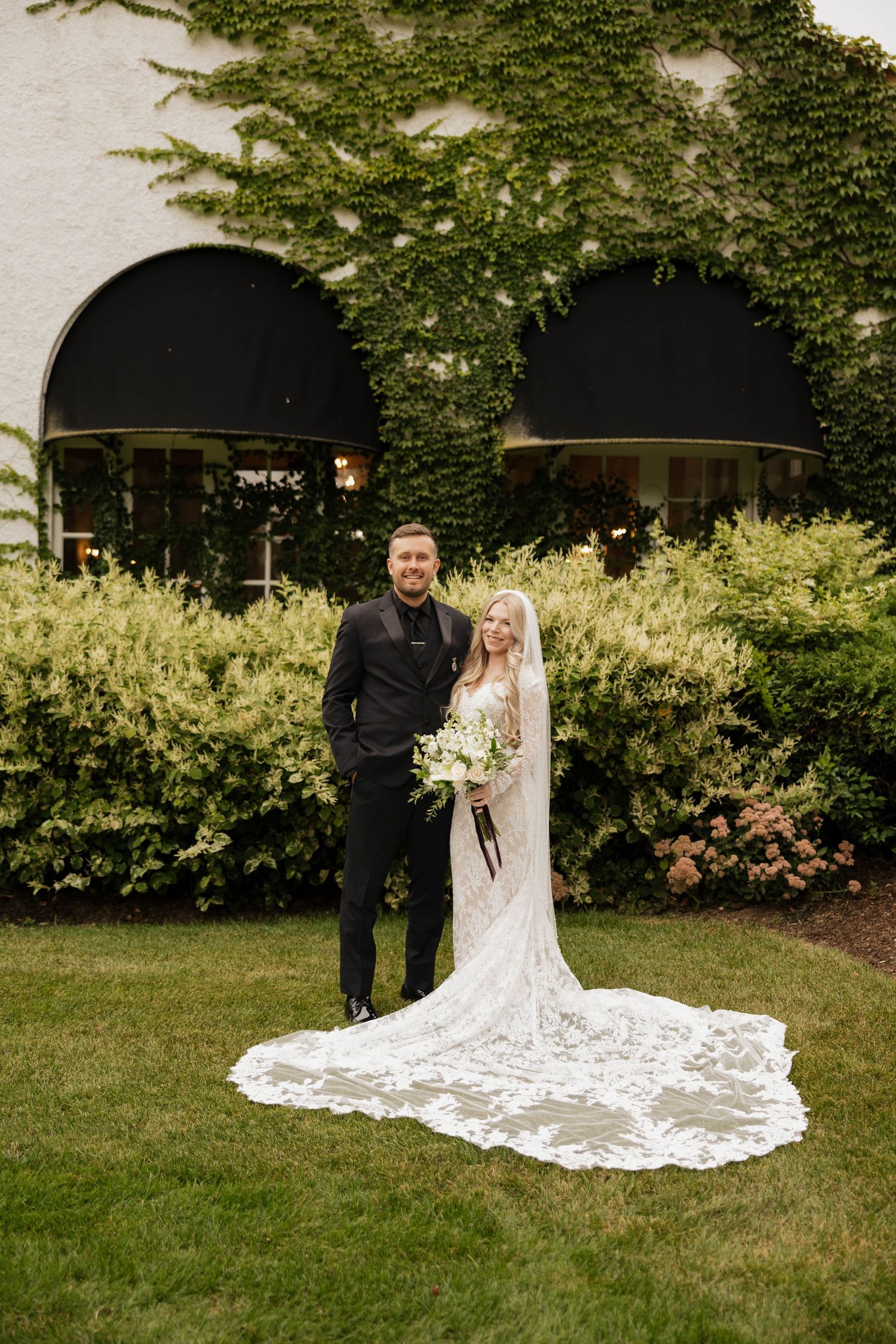 Bride in a lace gown with a long veil and groom in a black suit pose on a lawn in front of ivy-covered walls and blooming shrubs.