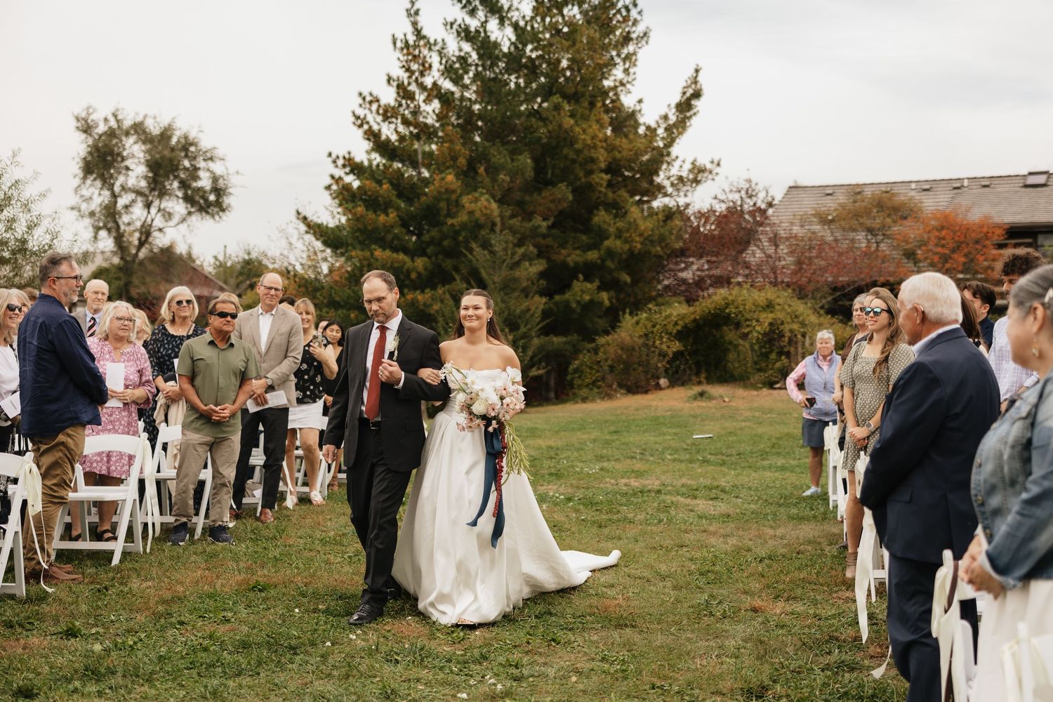 Bride in an off-the-shoulder gown walks down the grass aisle on her father's arm as wedding guests watch from white chairs.