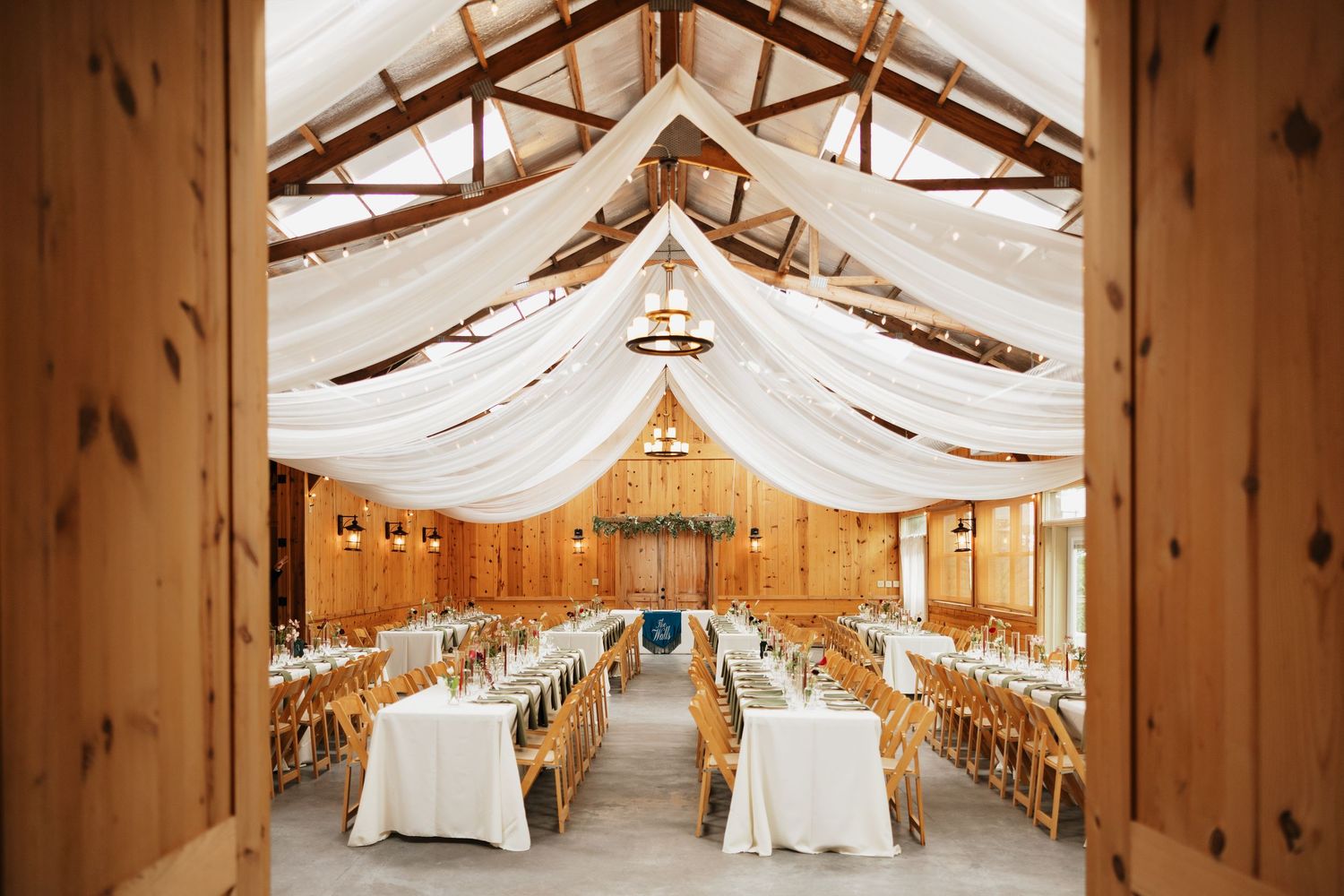Rustic wooden barn wedding venue with white draped ceiling fabric, long reception tables, and wooden chairs set for a celebration.