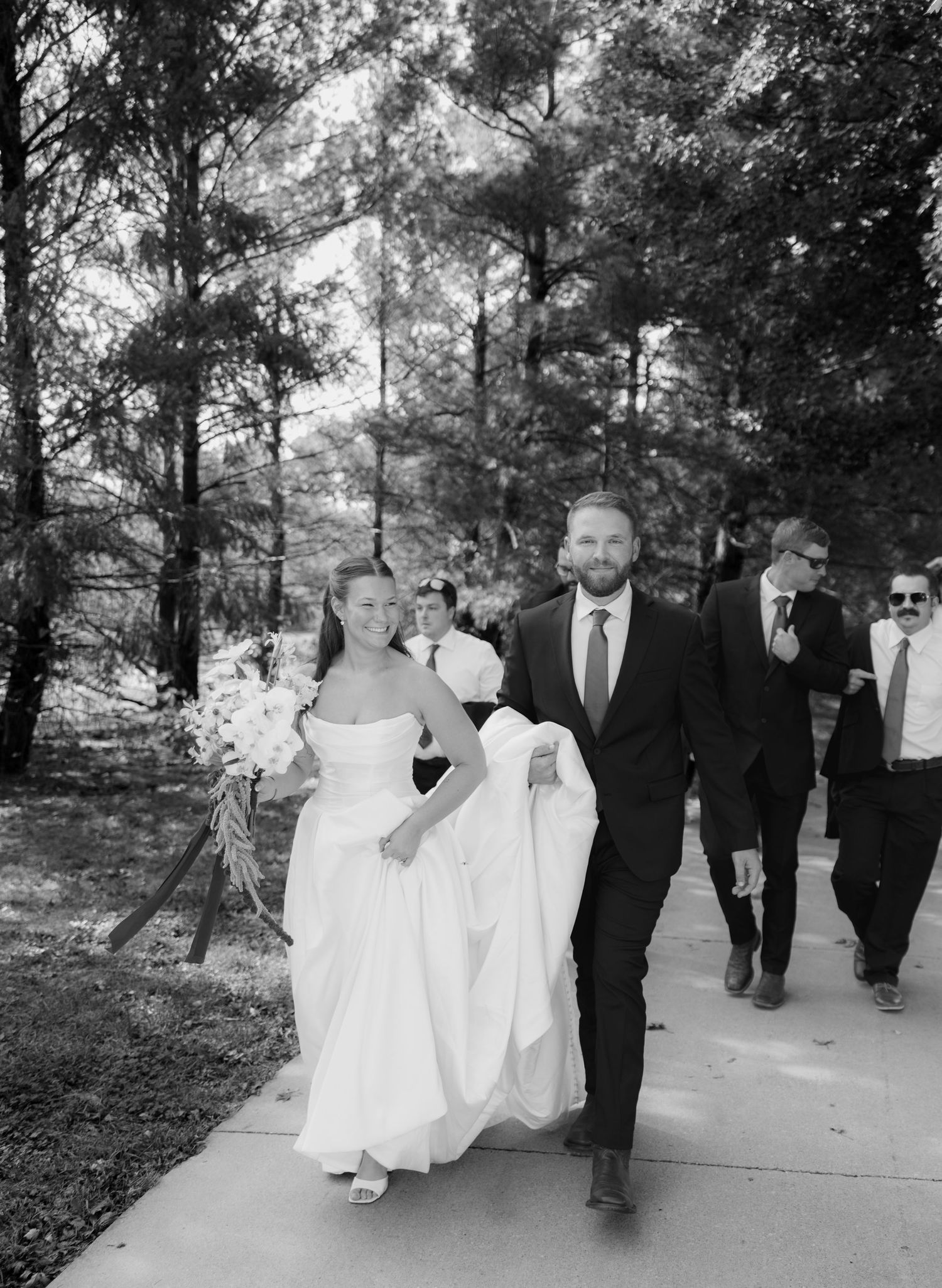 Black and white photo of a bride in a strapless gown holding a bouquet, walking beside the groom and wedding guests along a tree-lined path.