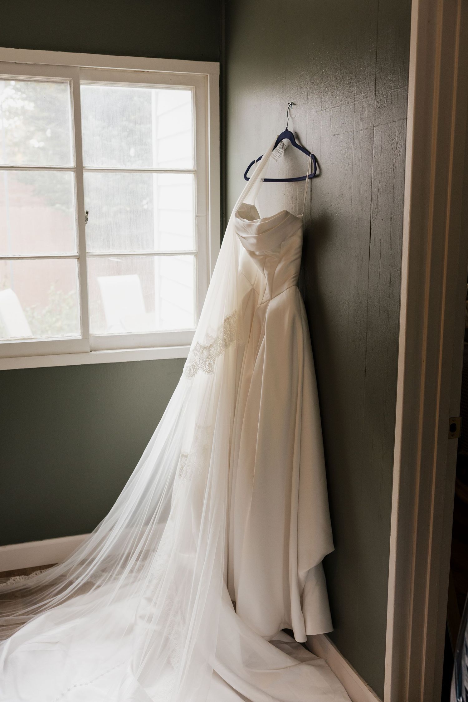 Strapless ivory wedding gown with a long flowing veil hangs on a hanger near a window in a softly lit green room.