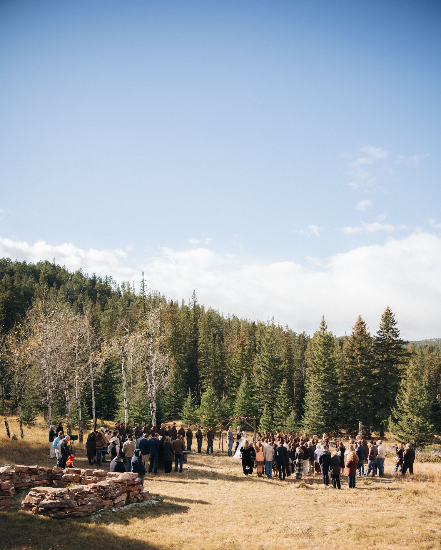 Large group gathered outdoors in a mountain setting surrounded by pine trees and clear blue sky.