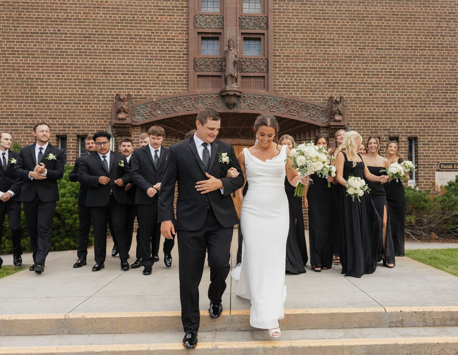 Wedding party members stand in front of a brick building wearing formal black and white attire.