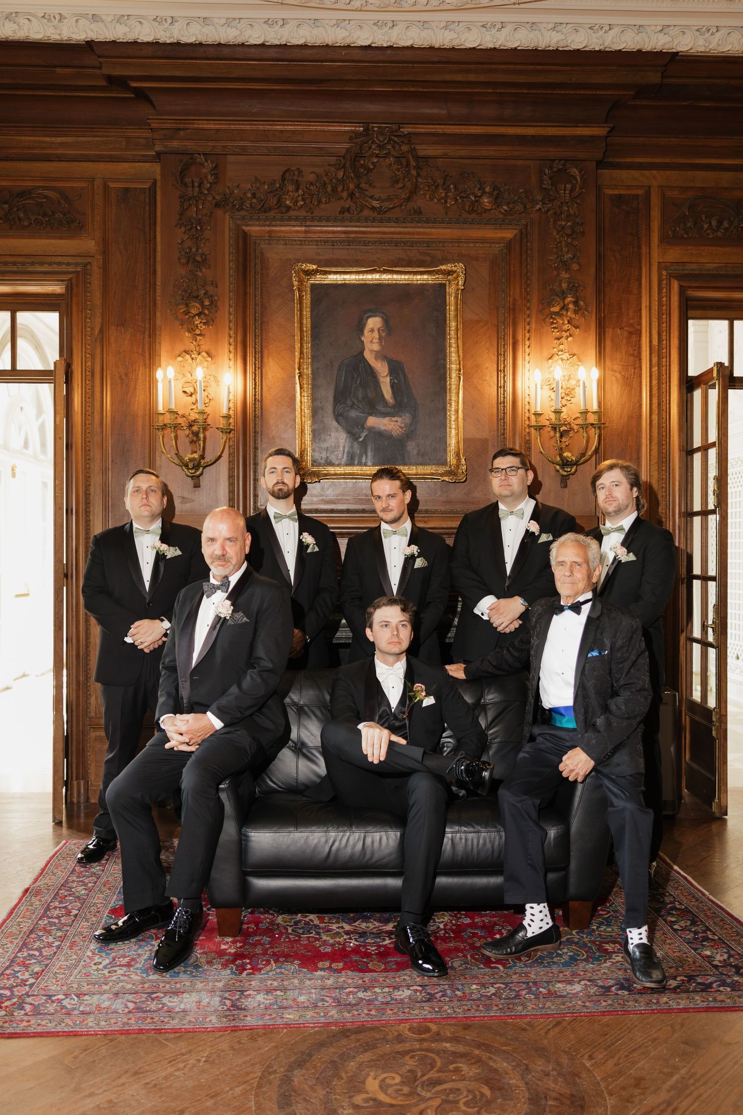 A group of nine groomsmen in black tuxedos pose in an elegant wood-paneled room with a portrait painting on the wall.