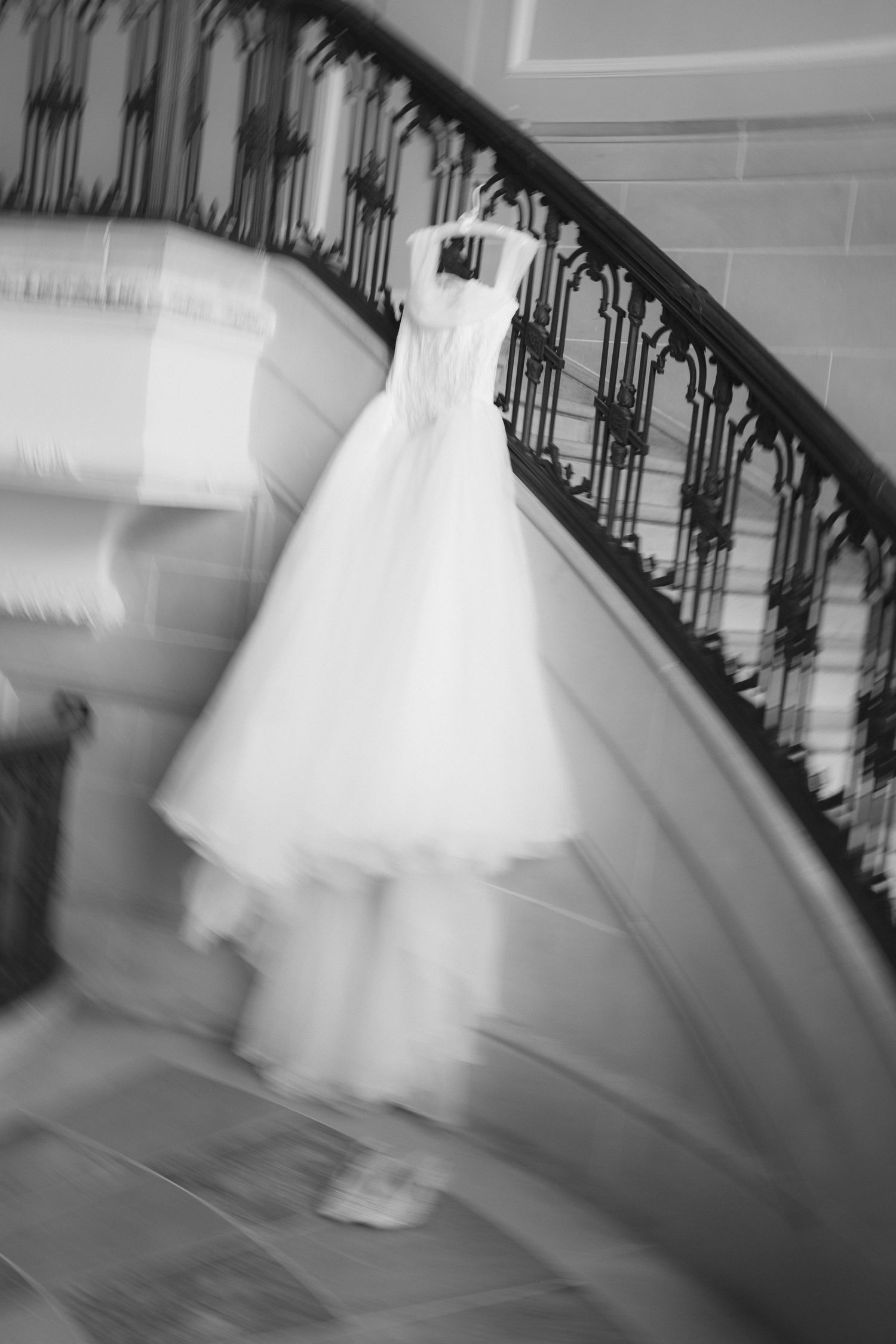 Black and white photo of an elegant wedding dress hanging on ornate spiral staircase railing in a grand venue.