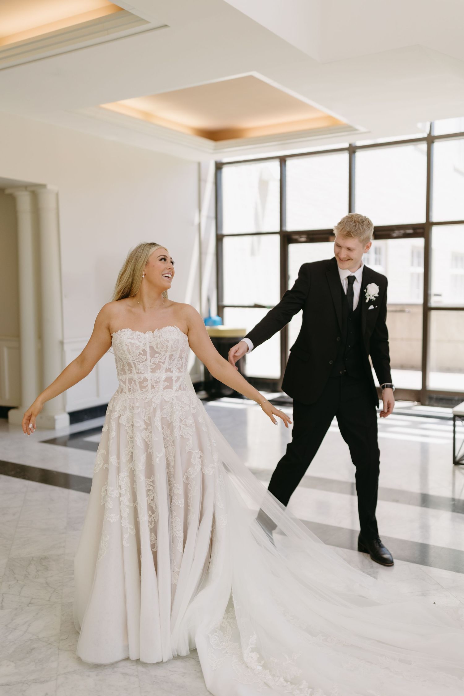 A couple in wedding attire dance together in a bright modern room with marble floors and large windows.