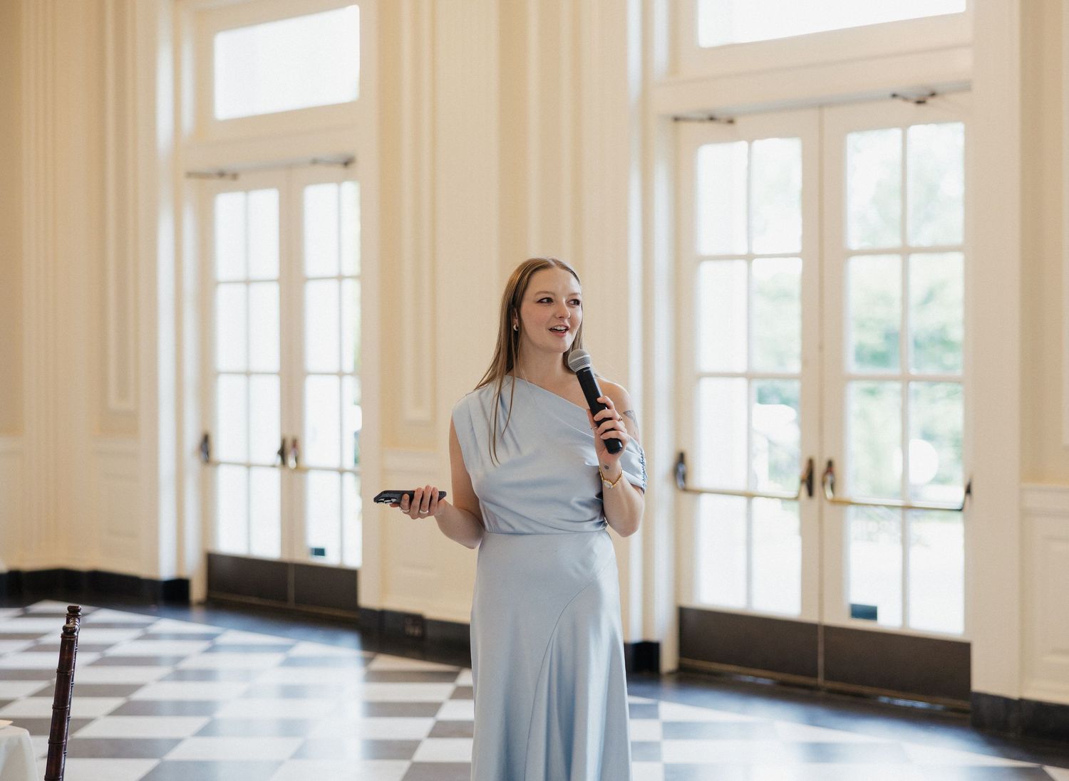 A woman in a flowing light blue dress stands gracefully in a bright sunlit room with checkered floors.