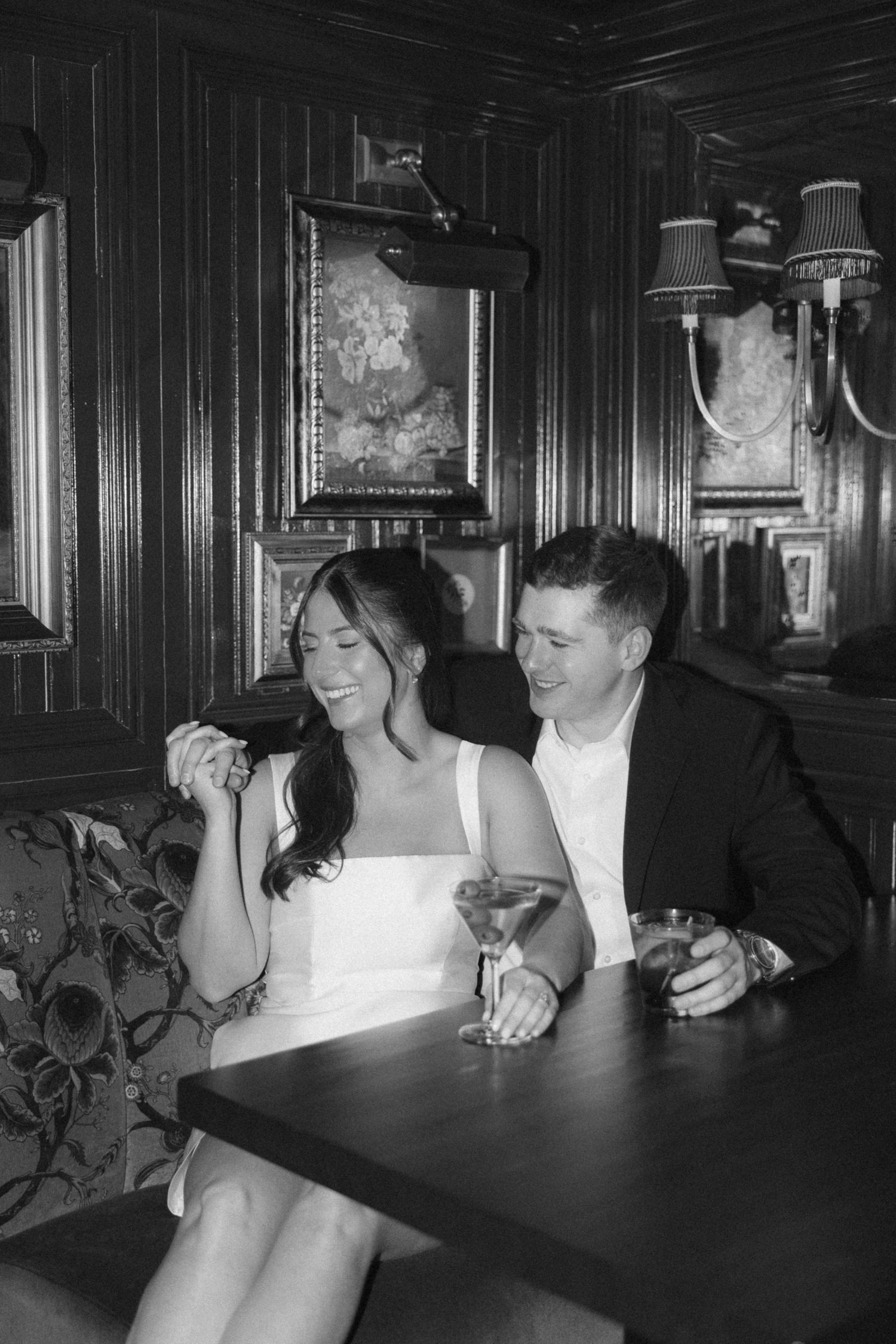 A couple in formal wedding attire laughs together while sitting at a dark wooden table in a dimly lit restaurant.
