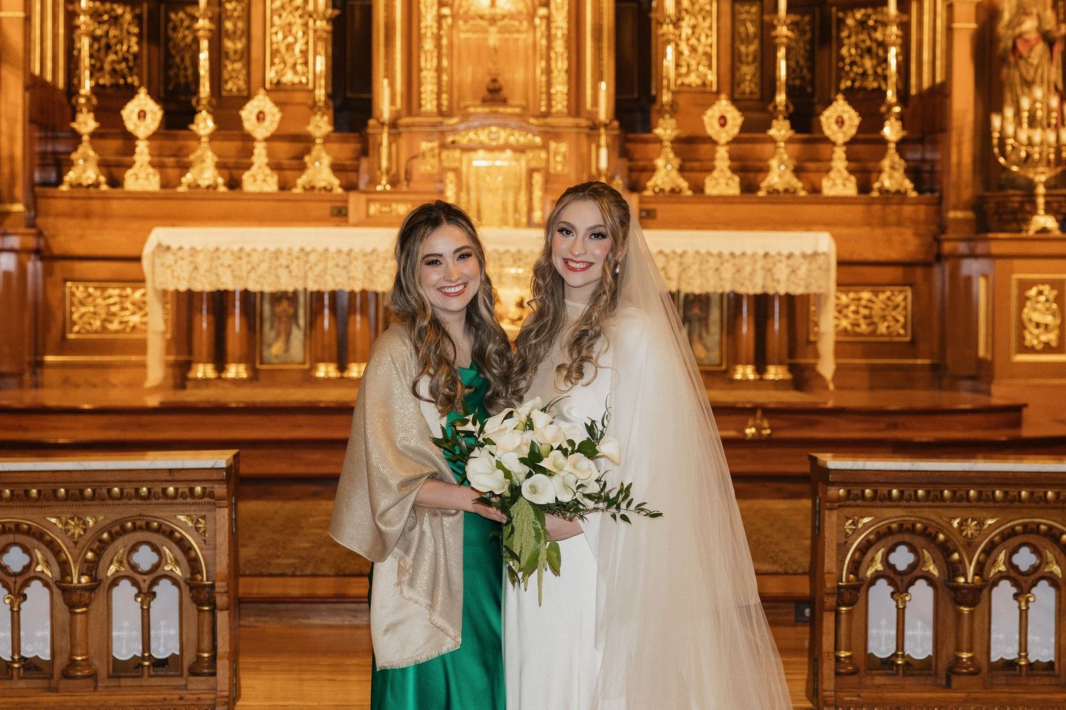 Two people stand at an ornate golden altar with one wearing a wedding veil and holding a white floral bouquet.