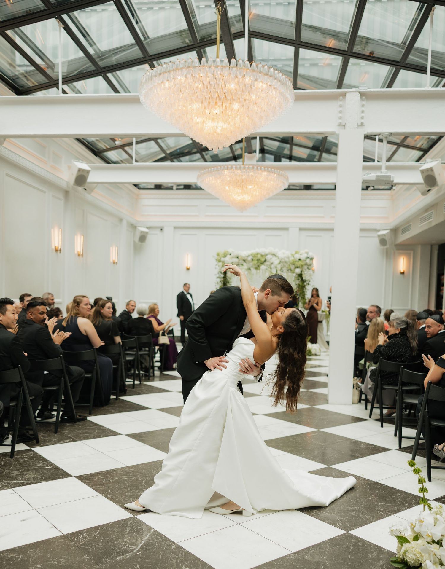 Bride and groom share a romantic dip kiss on black and white checkered floor under crystal chandeliers.