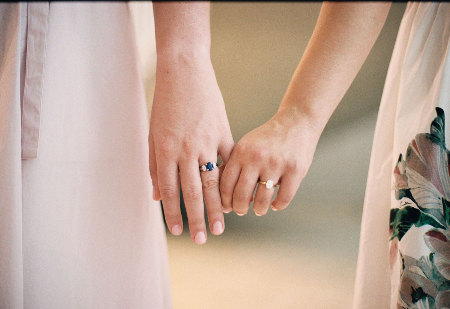 Two women holding hands, wearing elegant rings, dressed in pastel floral gowns, symbolizing love and commitment.