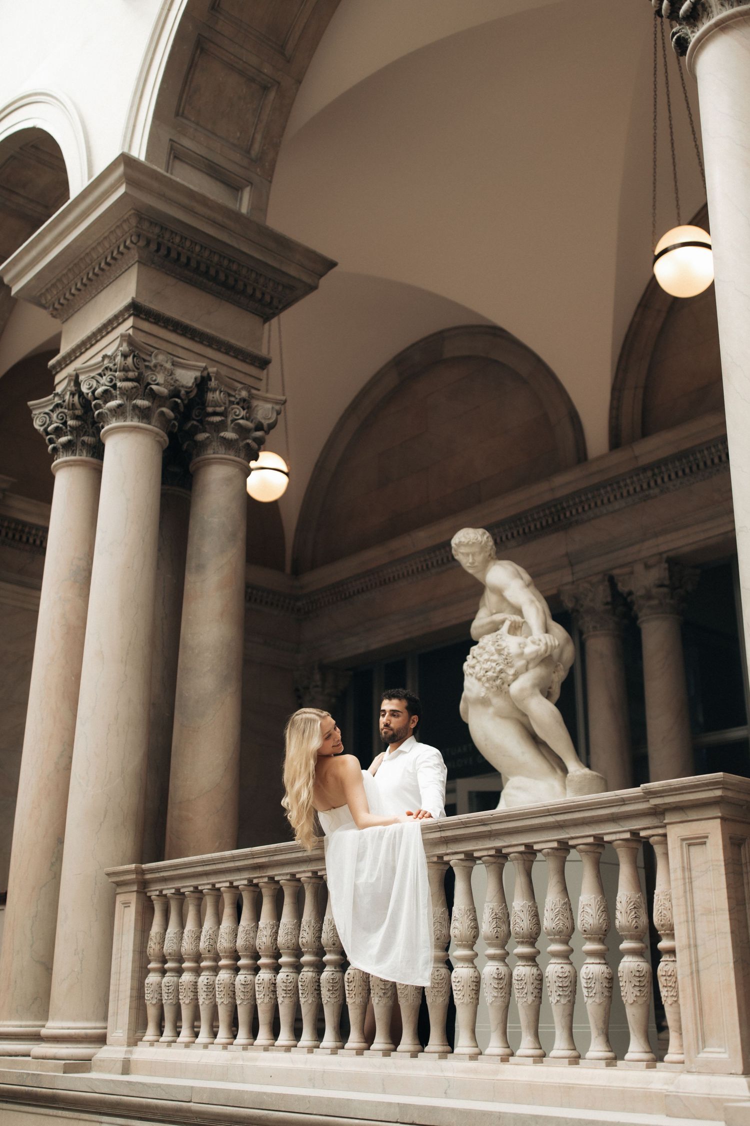 Couple in white attire standing by ornate balustrade in classical architecture with marble statue and columns.