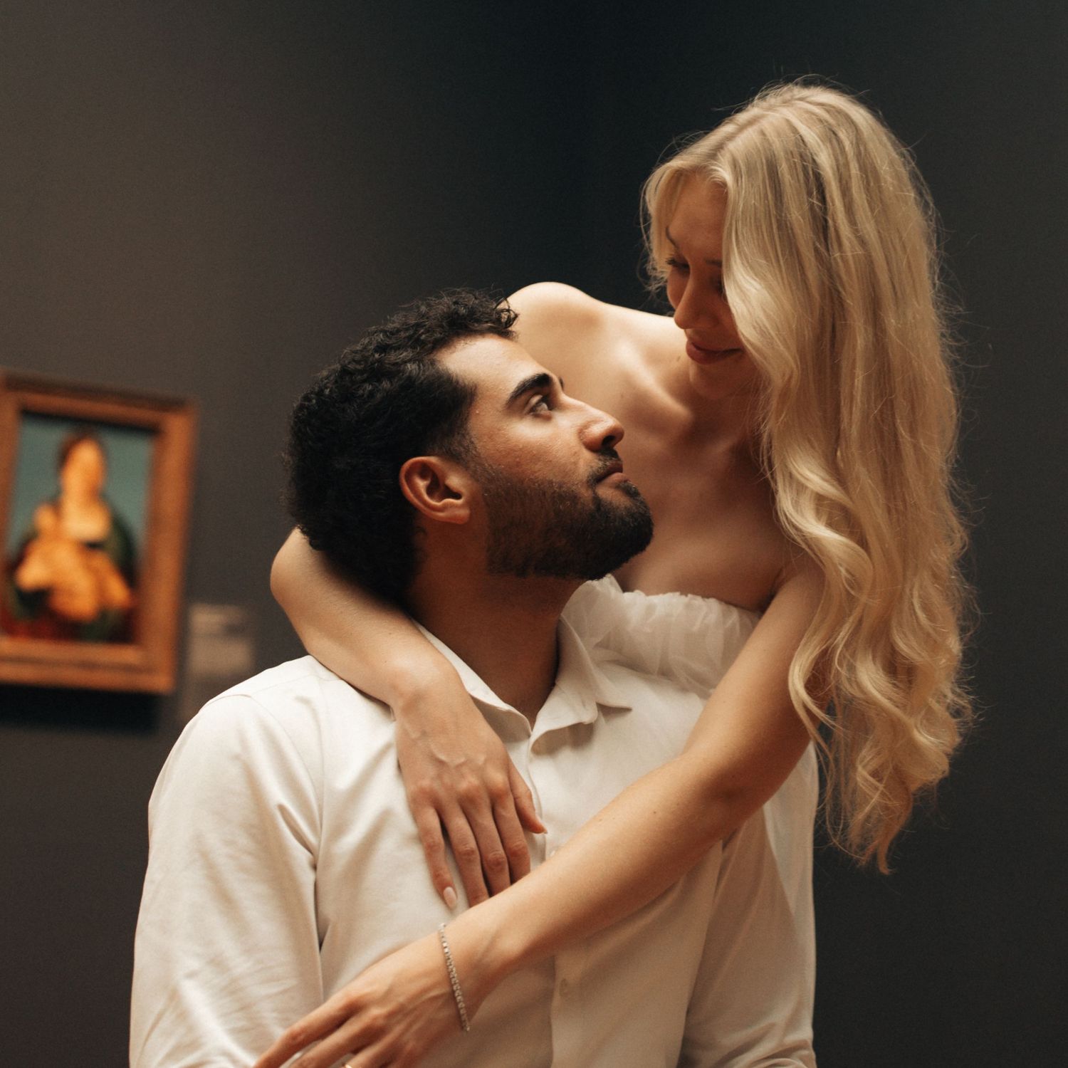 A couple shares a romantic moment in an art museum, with a painting visible on the wall behind them.
