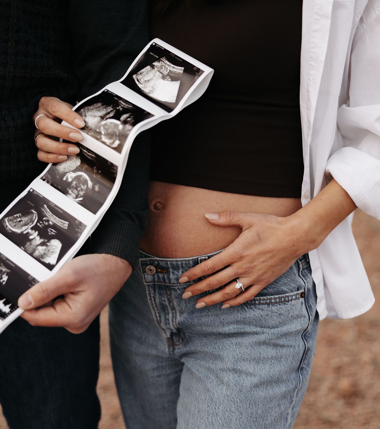 Pregnant woman in jeans holds ultrasound photos with partner touching her bare baby bump, wearing engagement ring.