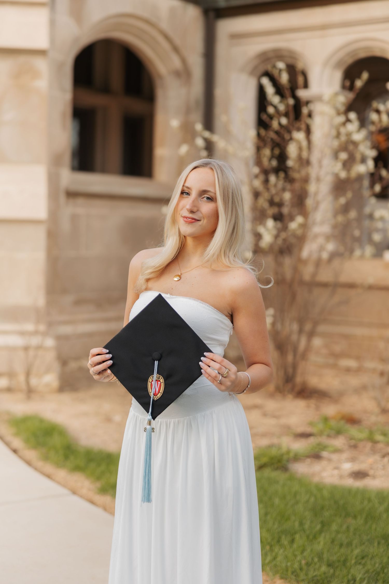 Blonde woman in white strapless dress holding black graduation cap with tassel outdoors near stone building.