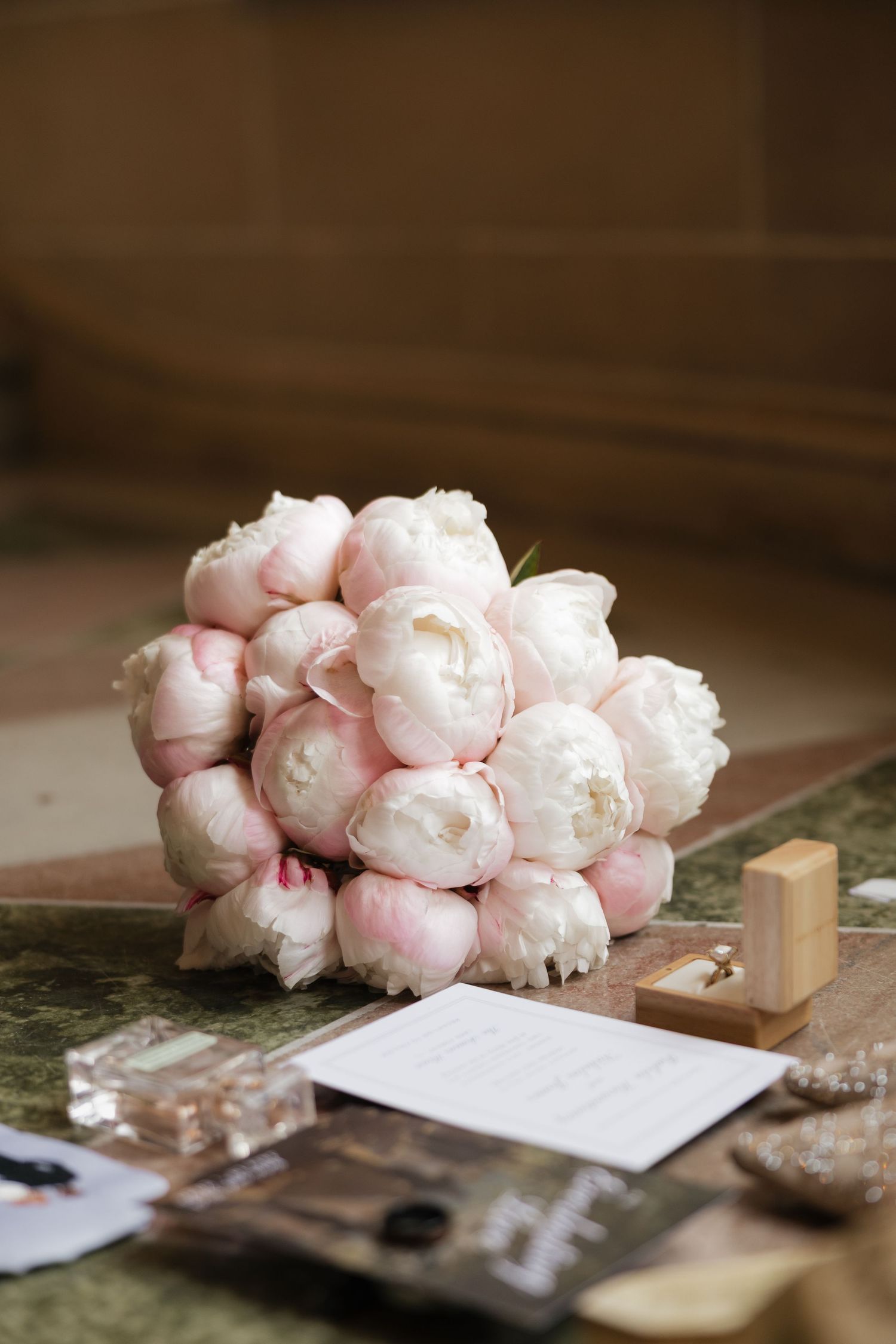 Elegant bridal bouquet of soft pink peonies on a marble table with wedding rings and cards nearby.