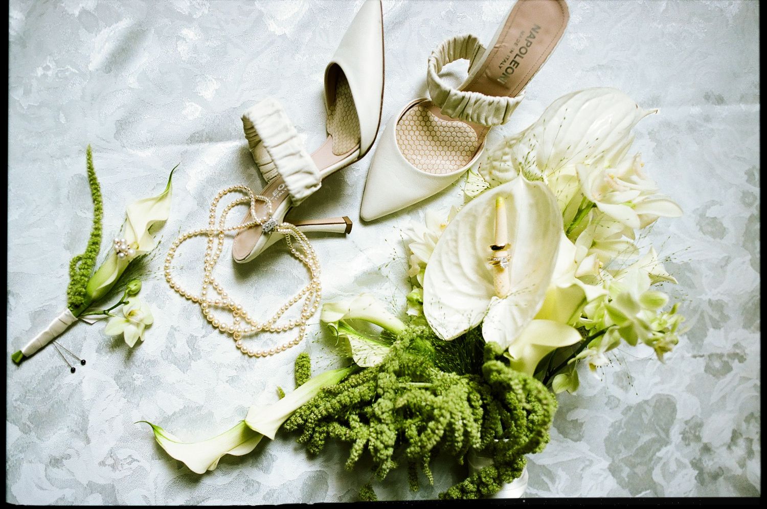 Elegant bridal accessories flat lay featuring ivory heels, pearl necklace, white calla lilies, and green boutonniere on marble.