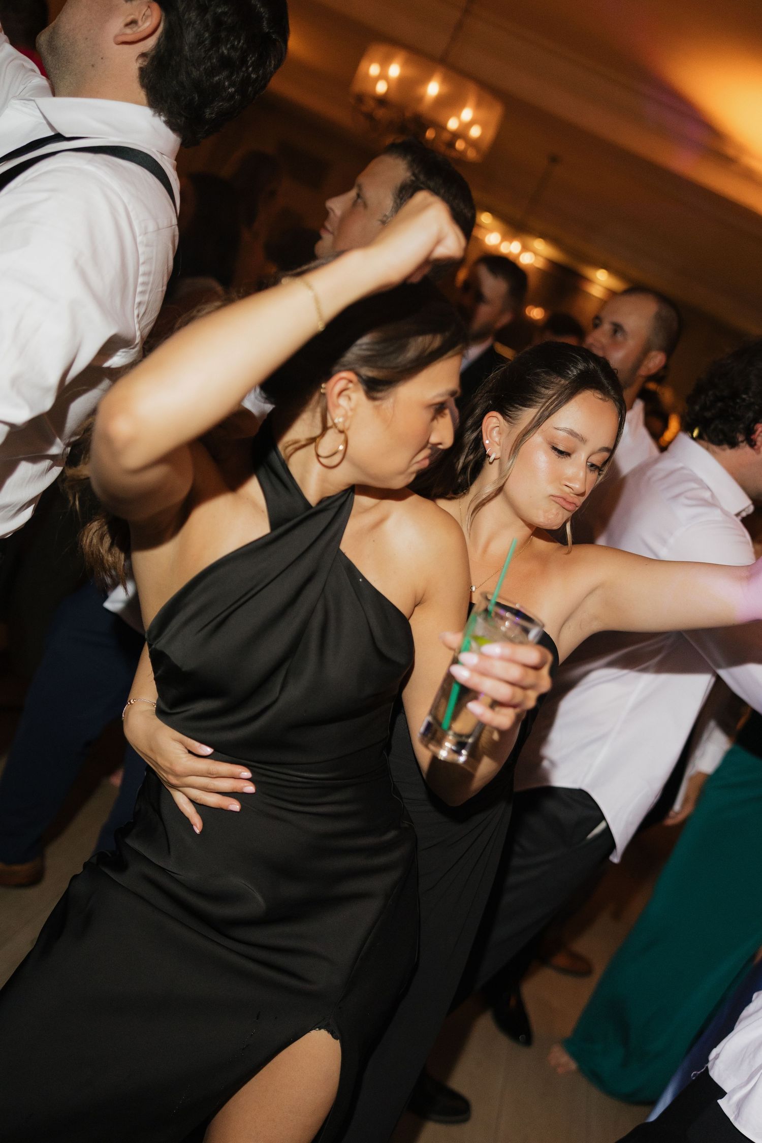 Guests dancing at a wedding reception, woman in black halter dress holding a cocktail on a crowded dance floor.