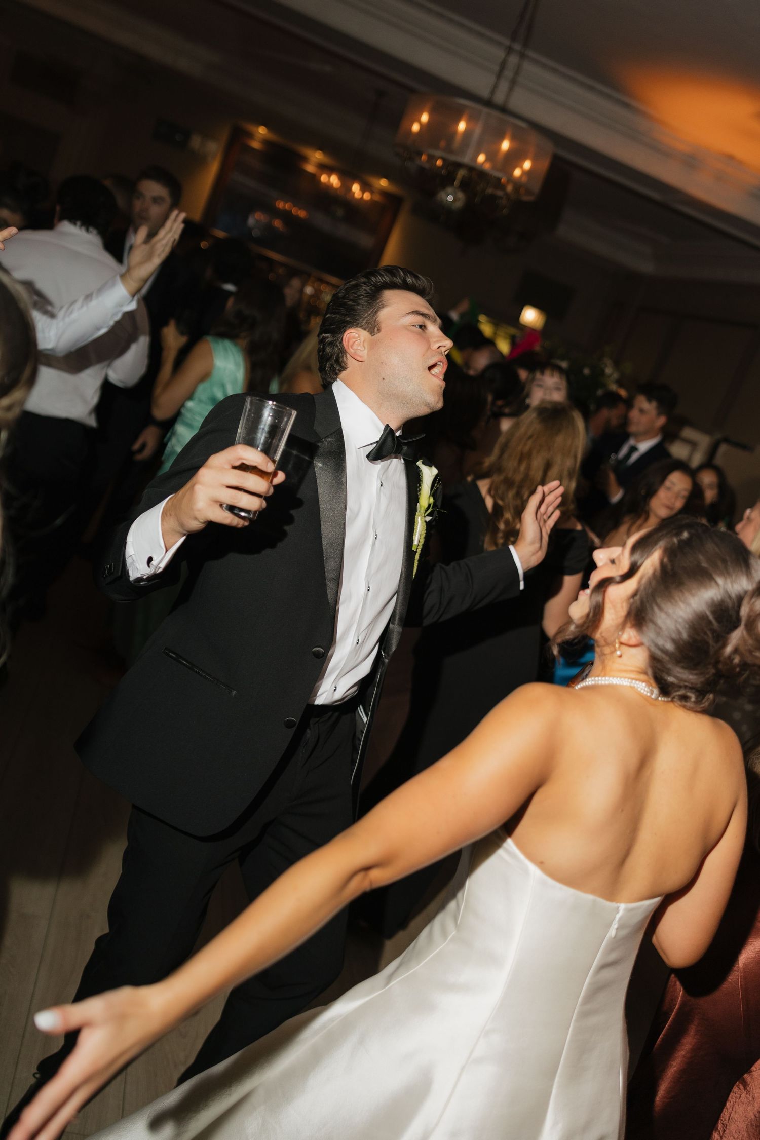 Bride and groom dancing joyfully at wedding reception, groom in tuxedo holding drink, bride in white gown.
