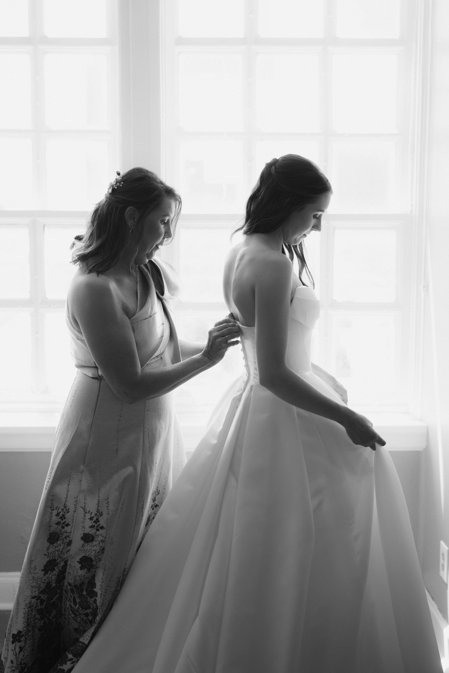 Black and white photo of bridesmaid helping bride button her wedding gown in front of a bright window.