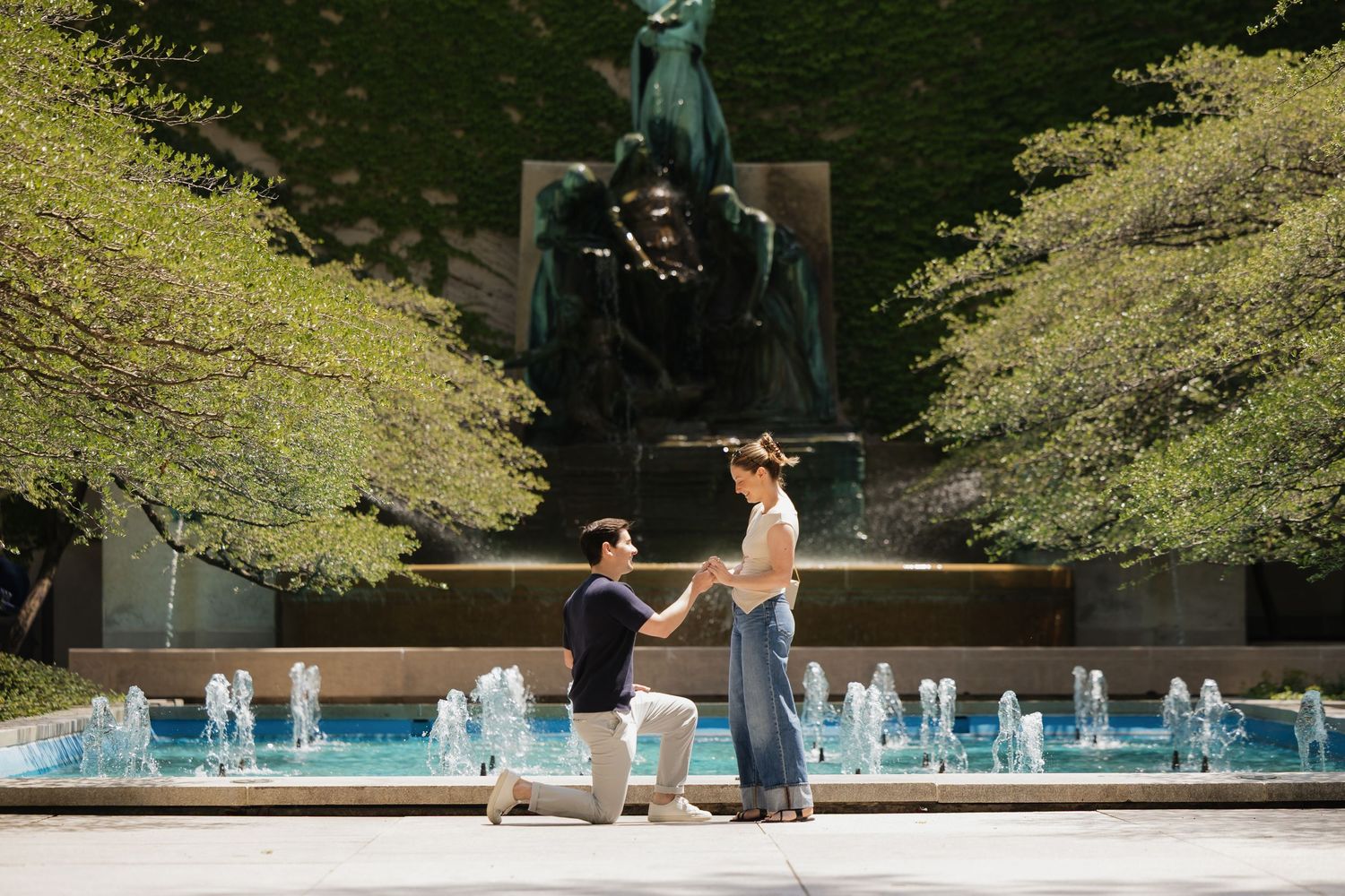 Man proposing to woman on one knee by a fountain with a bronze statue and lush green trees in background.