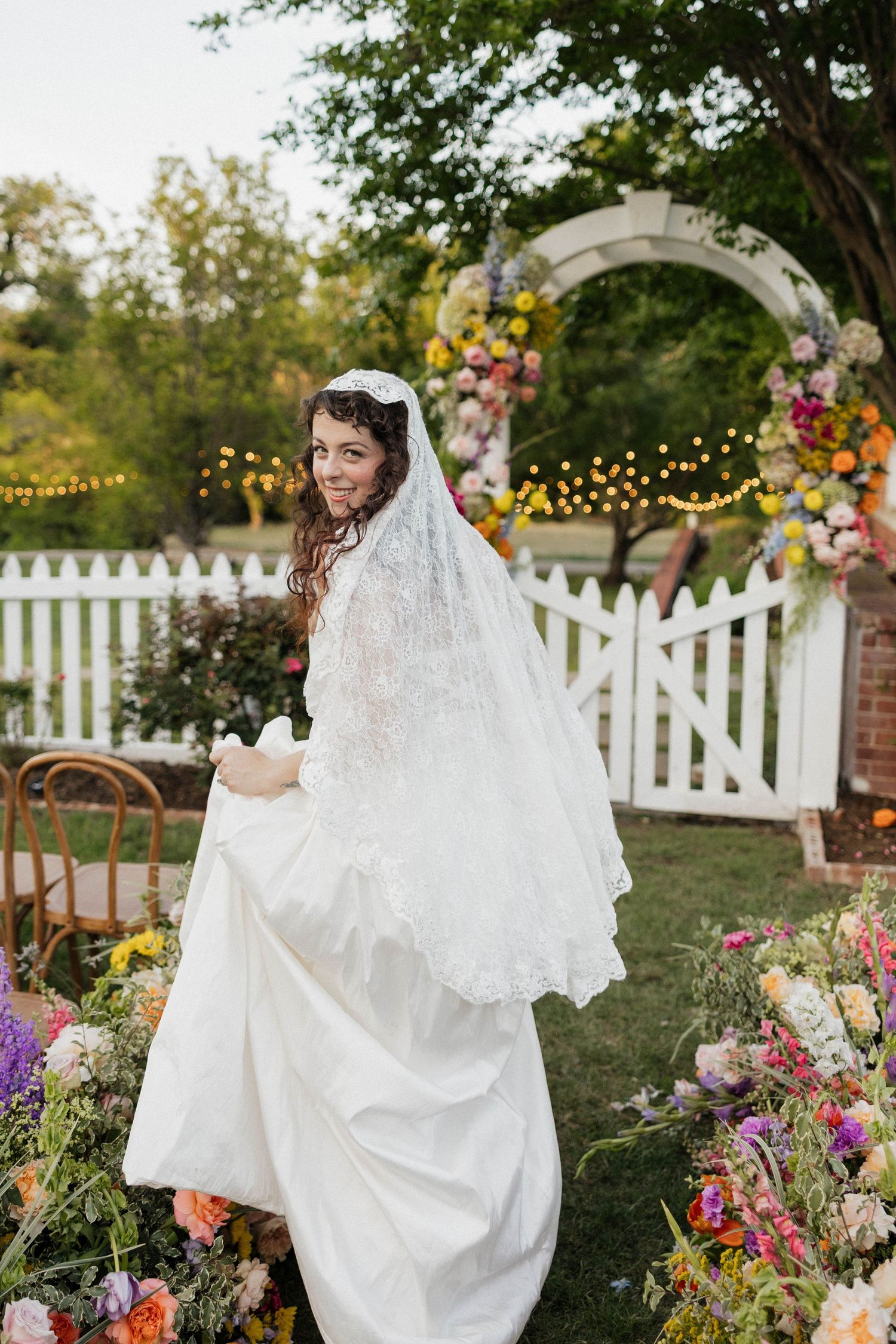 Smiling bride in white lace veil and gown holding dress train in colorful flower garden with white picket fence arch.