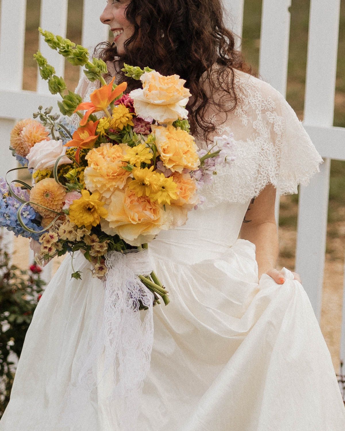 Bride in lace wedding dress holding a colorful bouquet of yellow, orange, and blue flowers outdoors.