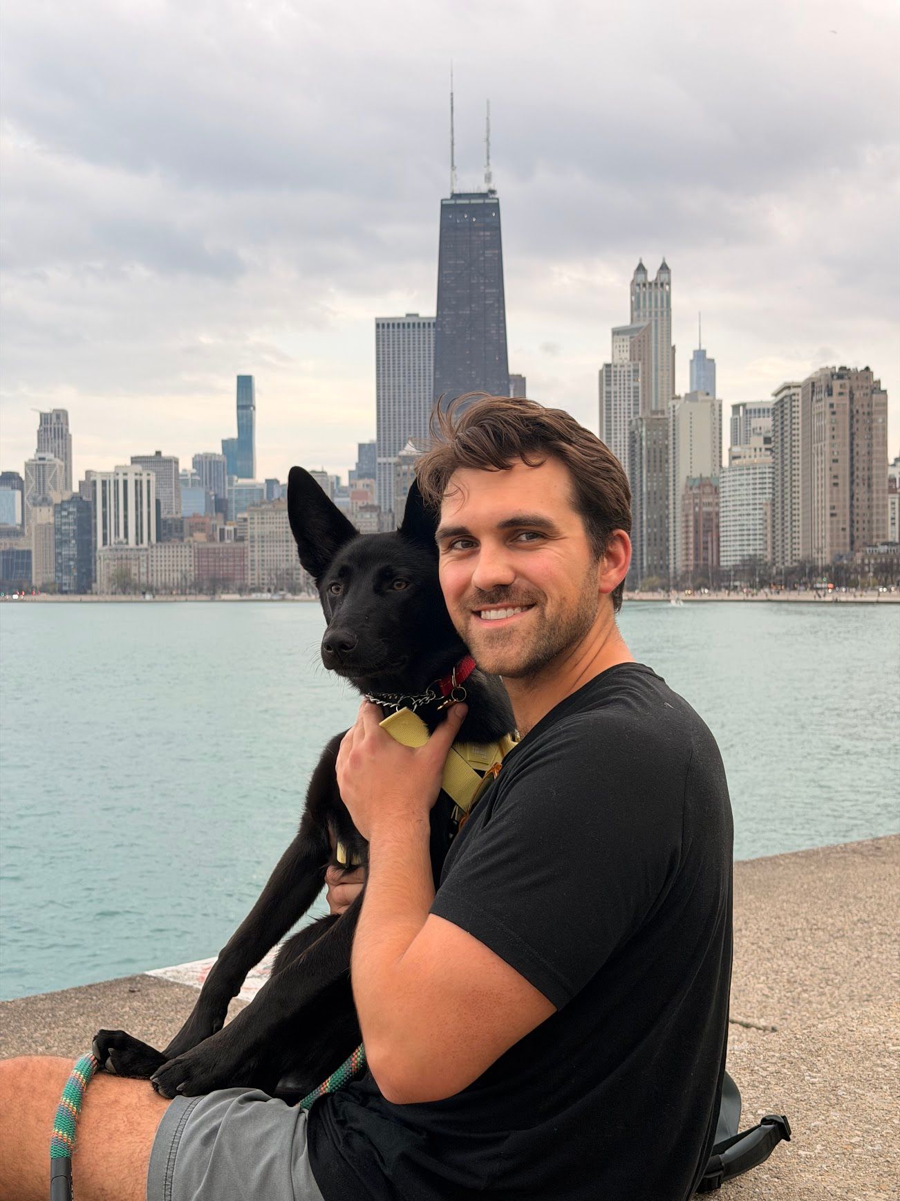 Man smiling with black dog on Chicago waterfront, John Hancock Center skyline in background on cloudy day.