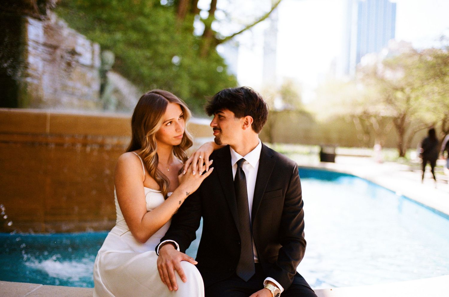 Bride and groom sitting together by a fountain, sharing a romantic moment in an urban park setting.