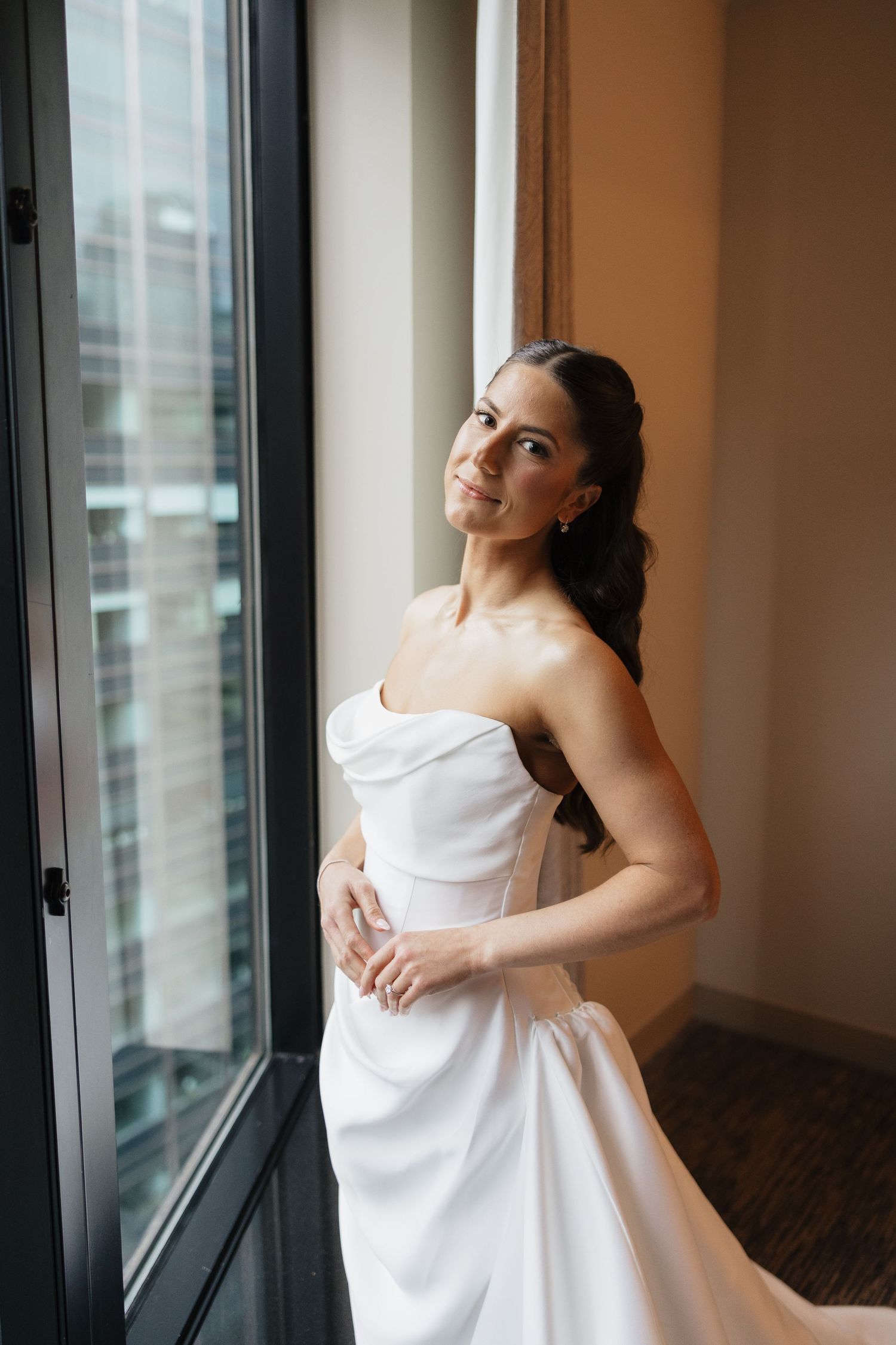 Elegant bride in strapless white wedding gown standing by a large window, smiling softly in a modern venue.