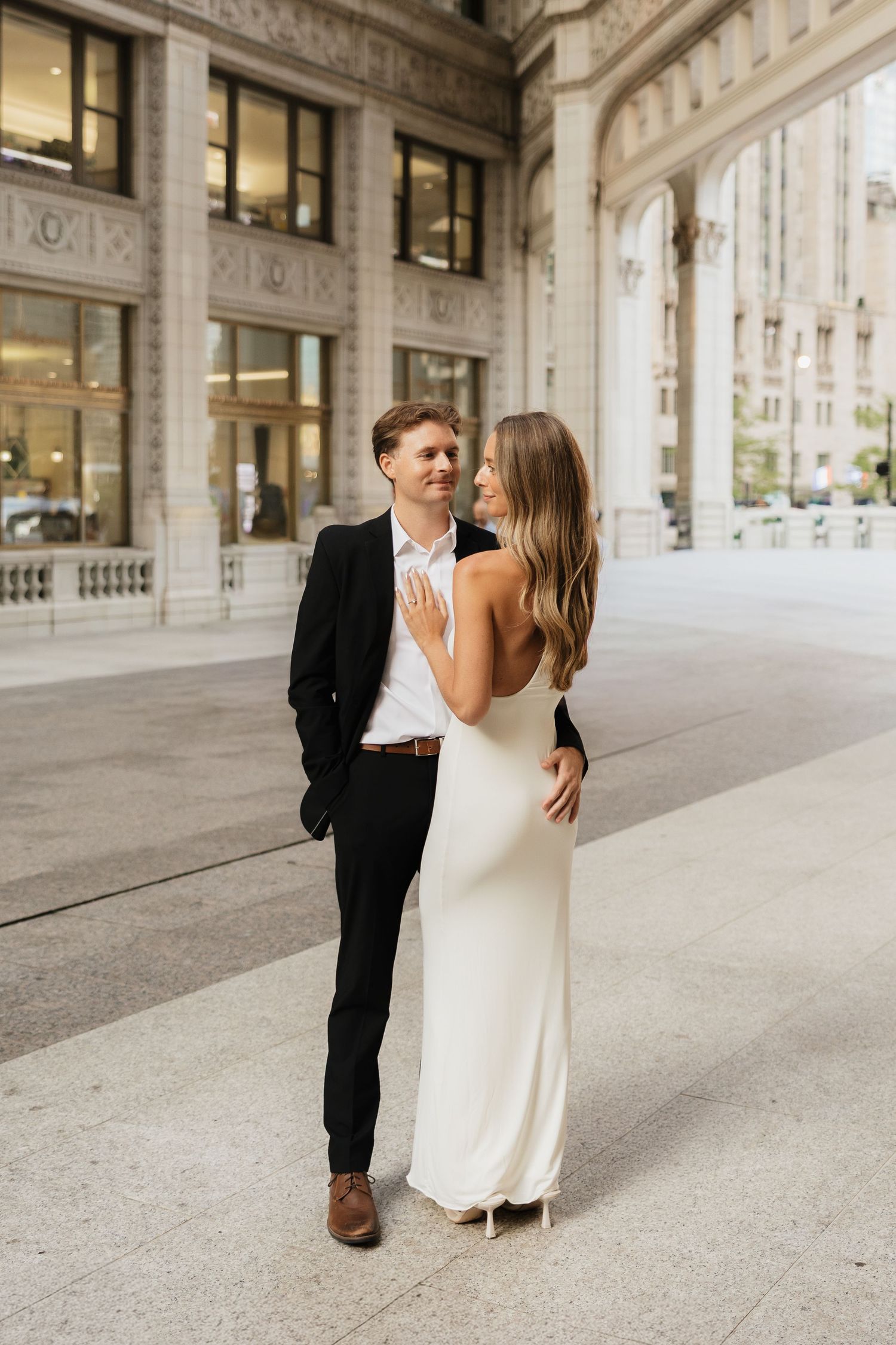 Elegant couple posing outside a classic building, woman in white gown, man in black suit, sharing a romantic moment.