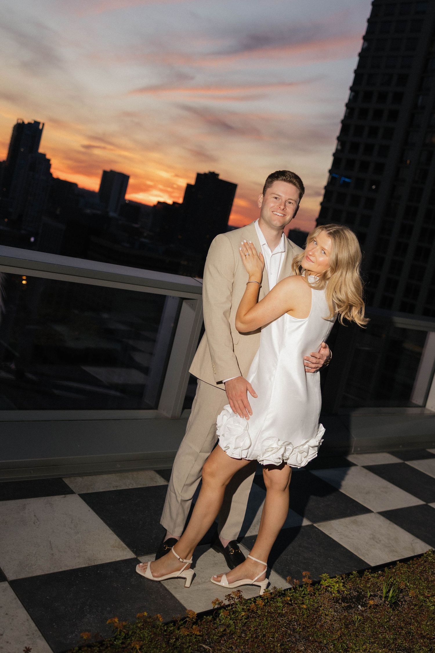 Couple embracing on a rooftop terrace with a stunning city sunset skyline behind them.