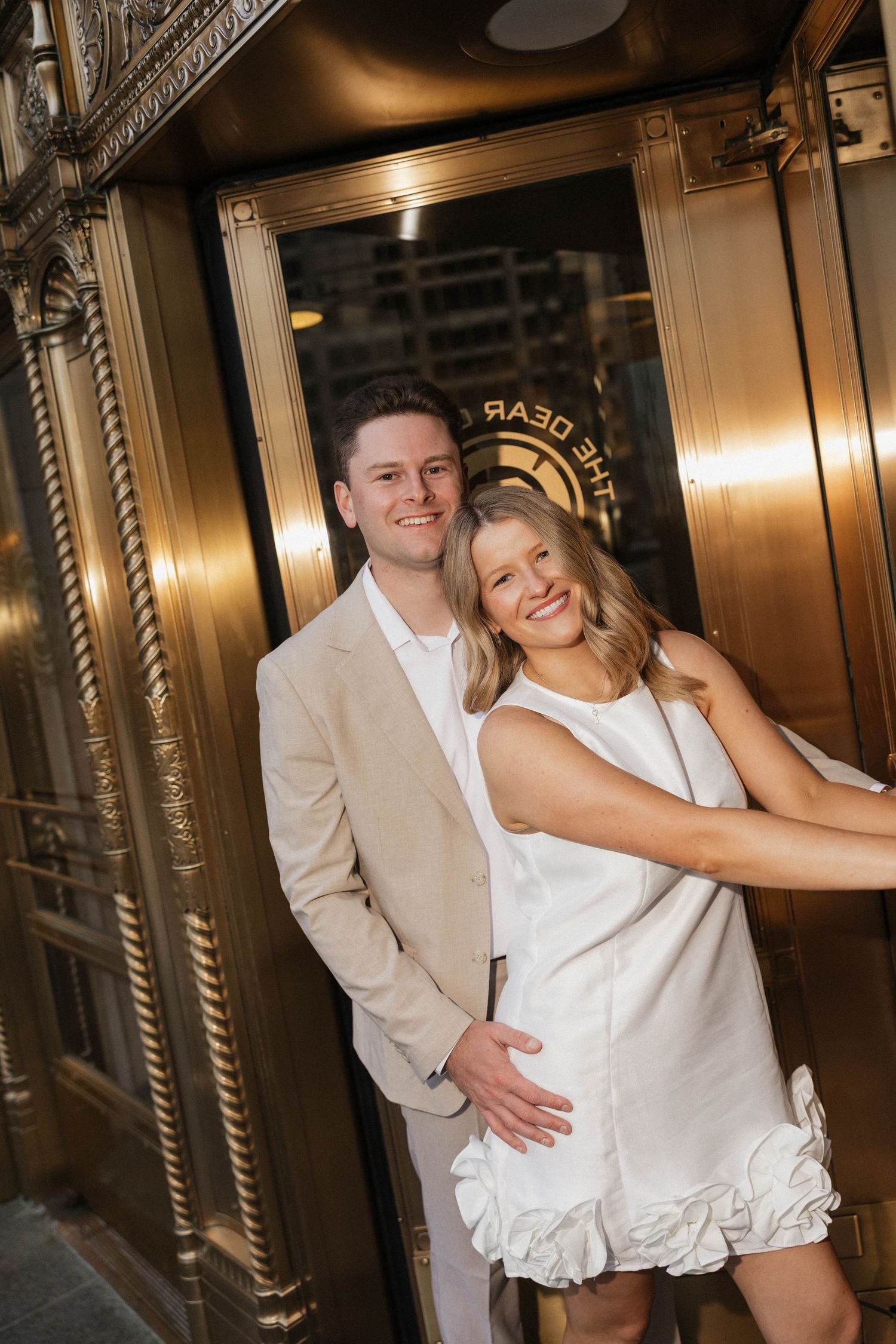 Smiling engaged couple posing at ornate gold revolving door entrance of elegant hotel.