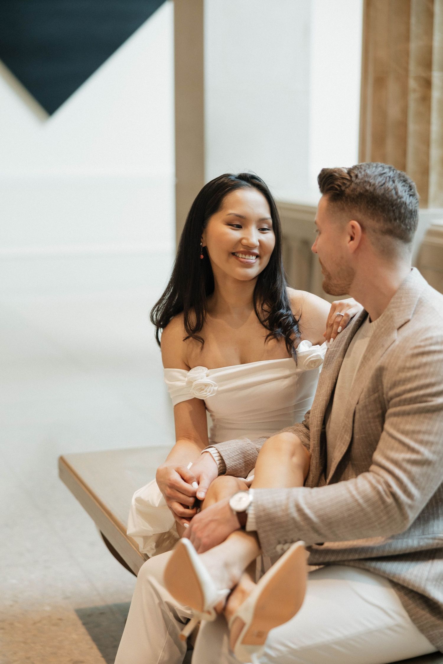 Smiling couple in elegant attire sitting together on a bench, woman in white off-shoulder dress holding hands with man in beige suit.