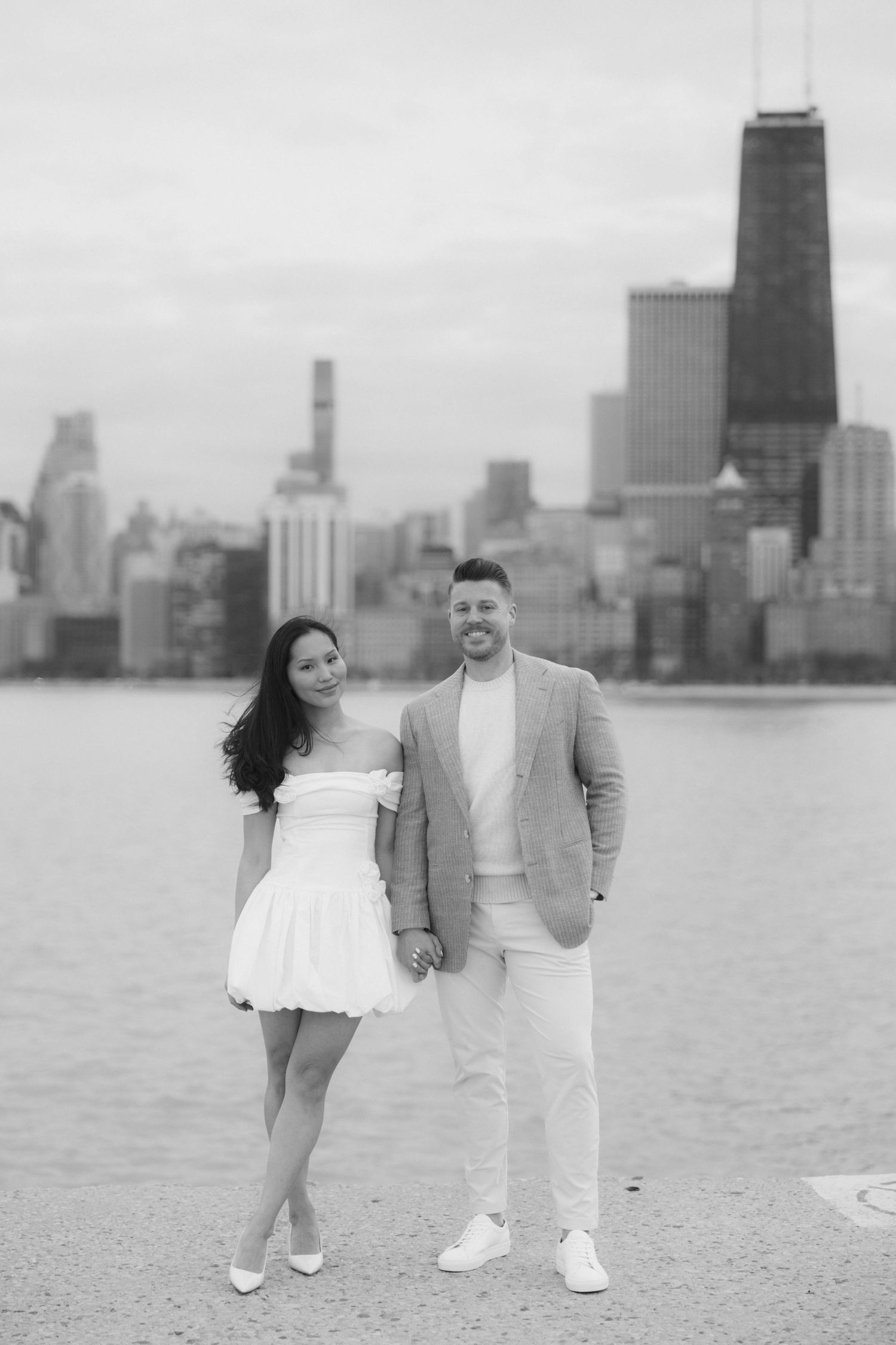 Black and white photo of a couple holding hands on Chicago lakefront with city skyline and John Hancock Center in background.