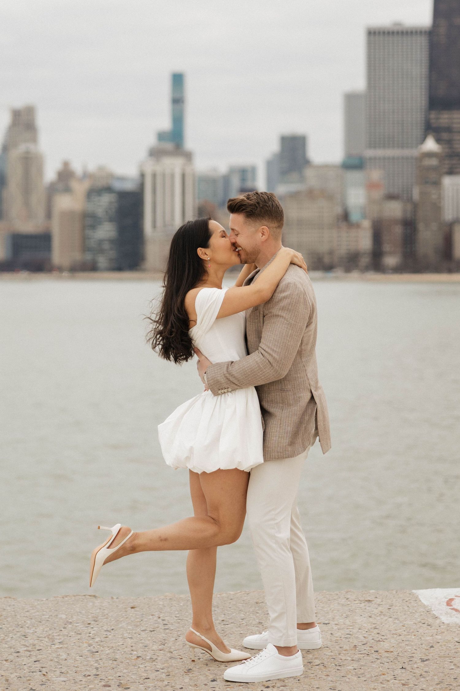 Couple sharing a romantic kiss by the waterfront with a city skyline backdrop.